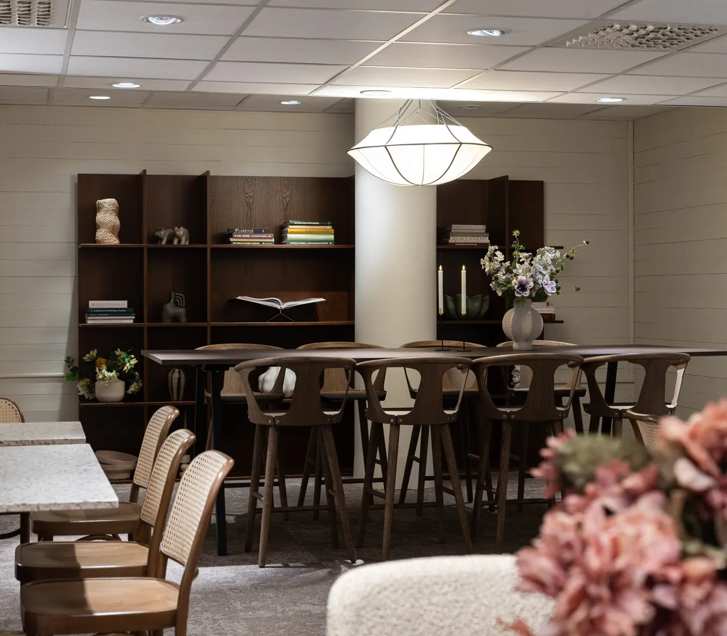 Modernt mötesrum med ett stort träbord och stilfulla stolar. Hyllor i bakgrunden dekorerade med böcker och prydnadsföremål. En elegant taklampa och en blombukett på bordet förstärker atmosfären