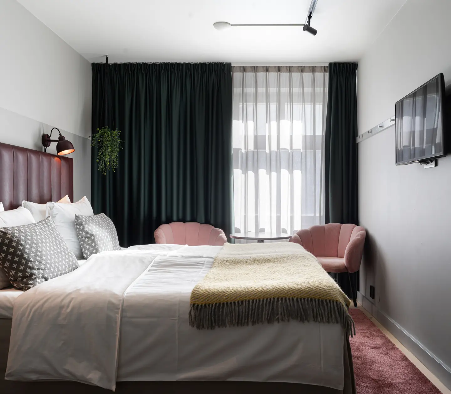 Modernt hotellrum med dubbelsäng, kuddar, rosa fåtöljer, fönster med mörka gardiner och väggmonterad TV