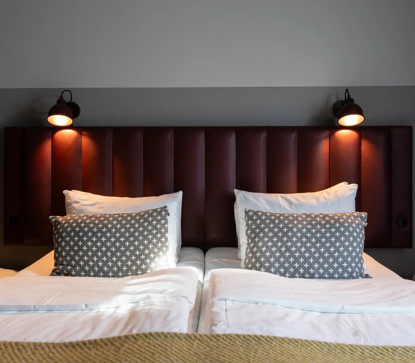 Hotellsäng med brun sänggavel, vita kuddar och grå mönstrade prydnadskuddar, upplyst av vägglampor