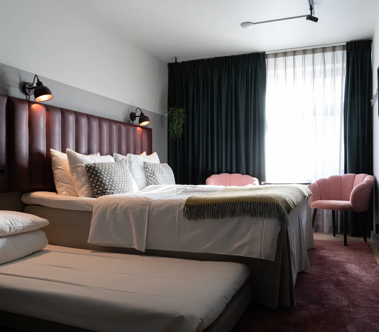 Modernt hotellrum med dubbelsäng, vit sängkläder och dekorativa kuddar. Mörka gardiner och rosa fåtöljer skapar en stilren atmosfär