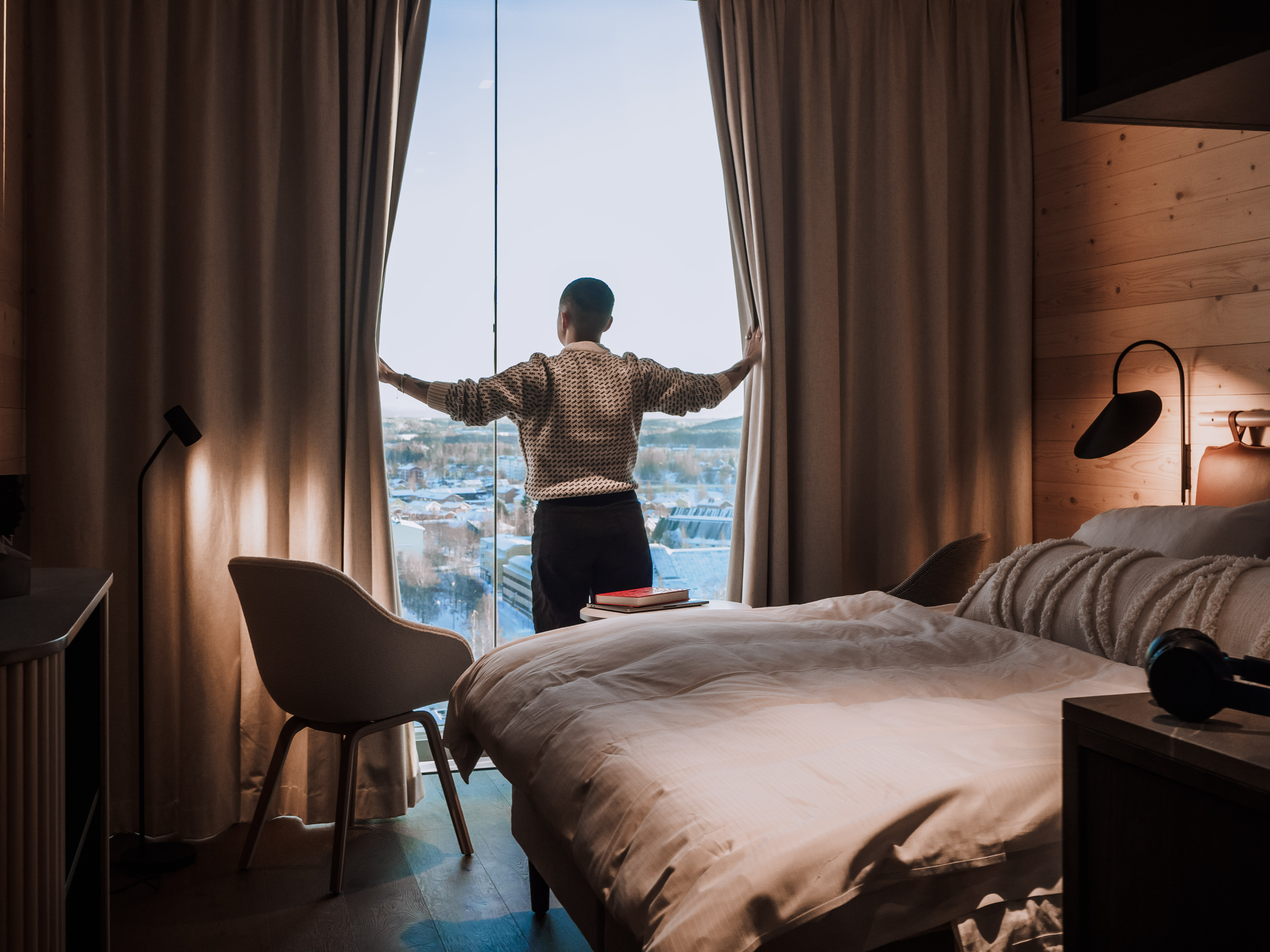 Person opening curtains in cozy hotel room with city view, featuring modern chair and plush bed in warm lighting.