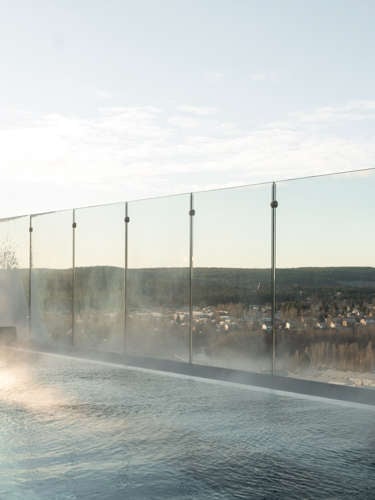 Outdoor pool with steam above the water surface, glass railing and view of Skellefteå