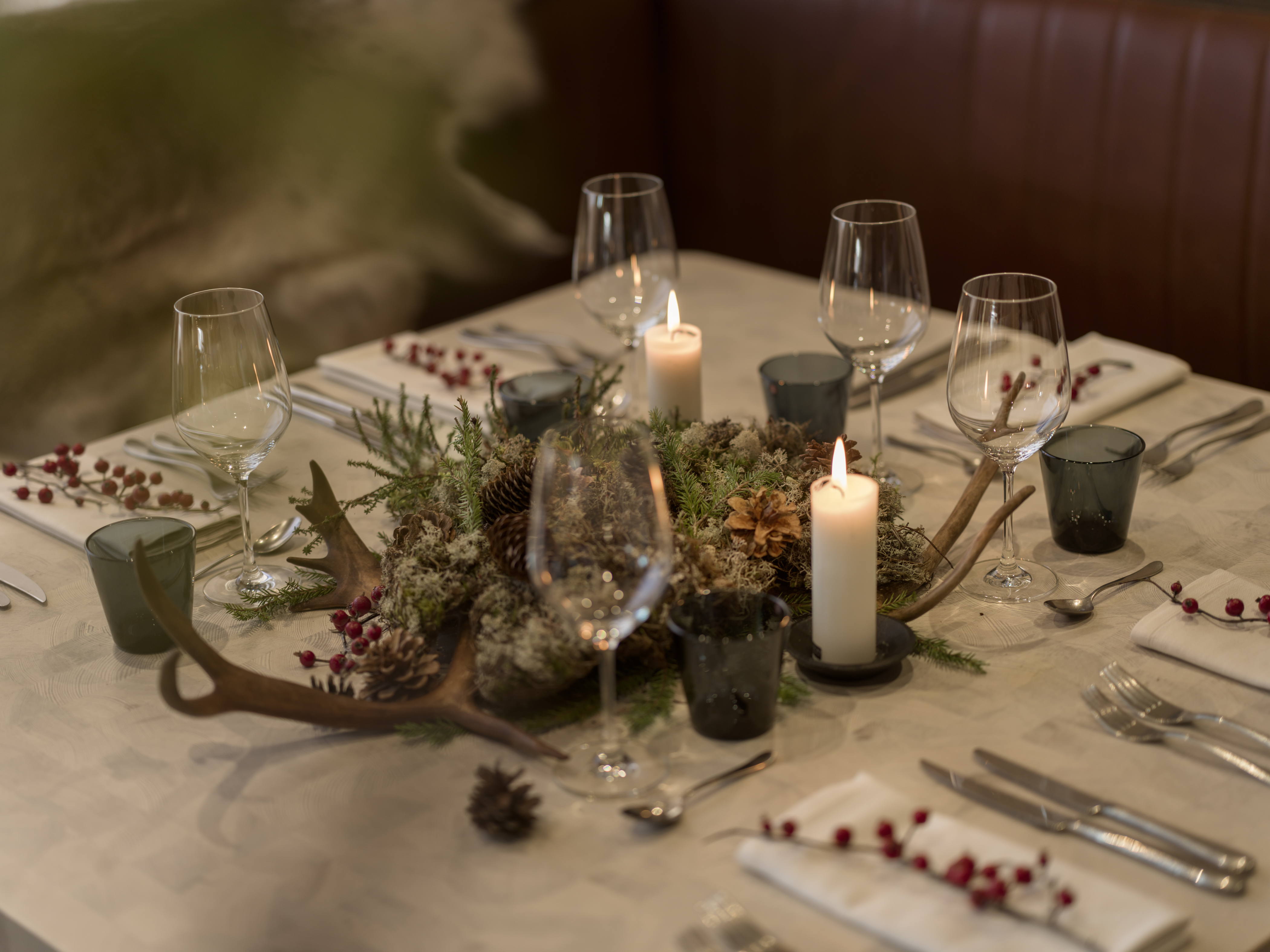 Elegant holiday table setting with candles, wine glasses, and rustic decorations featuring greenery and antlers.