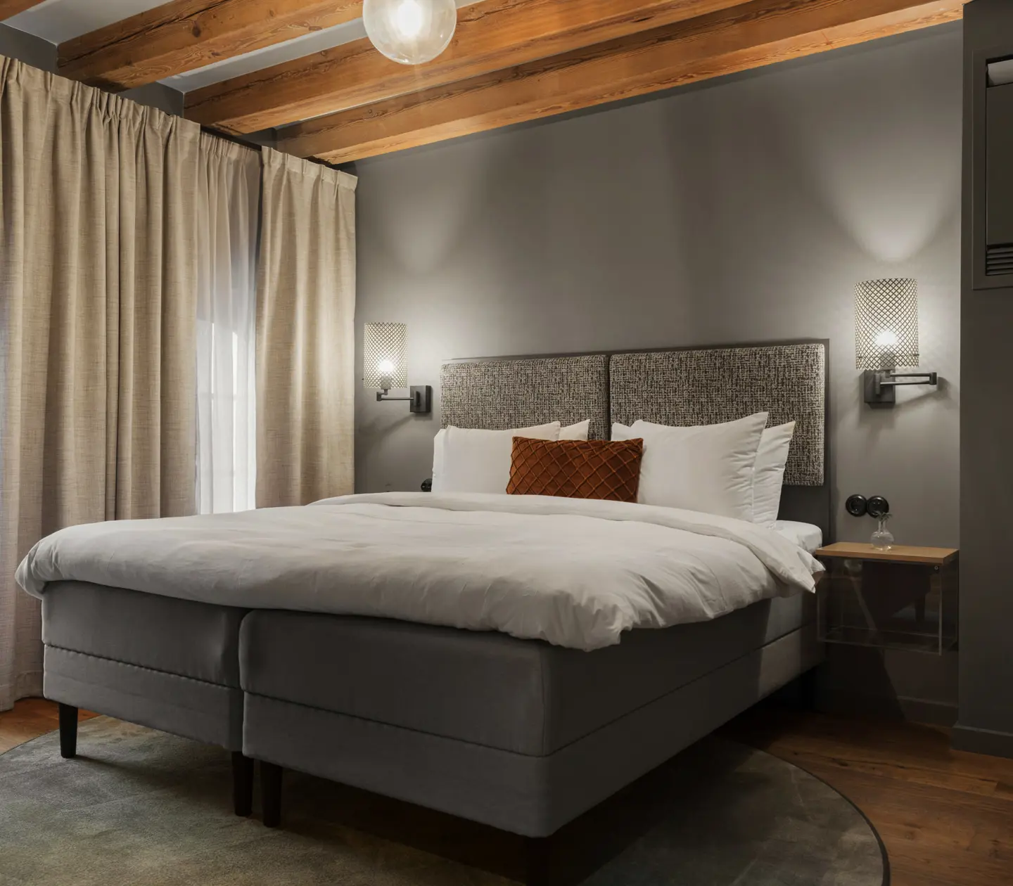 Ljust hotellrum med säng, beiga gardiner och träbalkar i taket