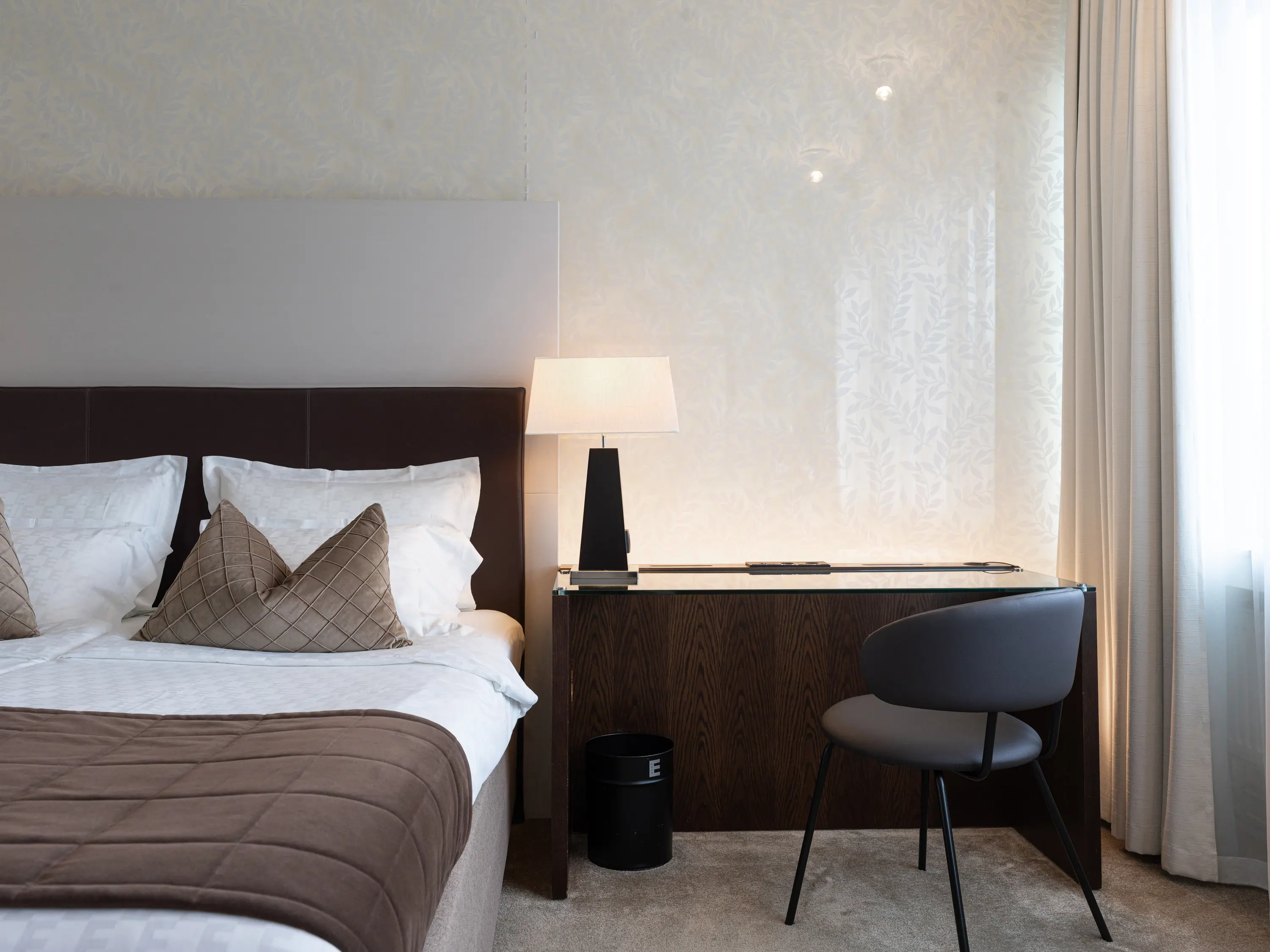 Modernt hotellrum med dubbelsäng, bruna överkast, skrivbord och stol, samt bordslampa vid sidan.