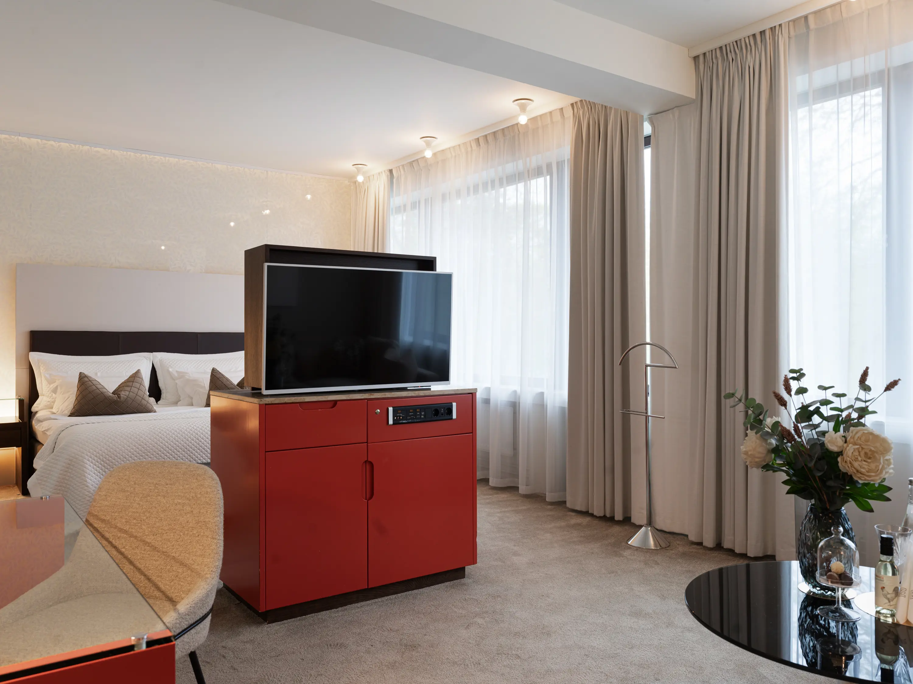 Lyxigt hotellrum med modern inredning, stor TV, bekväm dubbelsäng och blombuketter på ett glasbord. Naturligt ljus strömmar in genom stora fönster med vita gardiner.