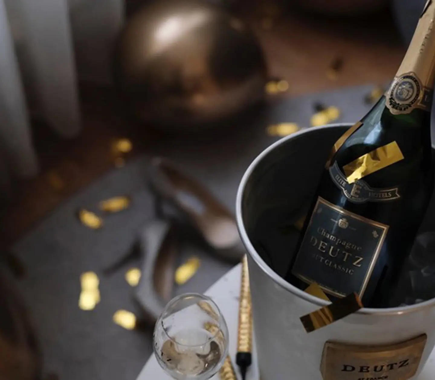 A bottle of Deutz Champagne in a silver ice bucket surrounded by gold confetti and a celebratory atmosphere.