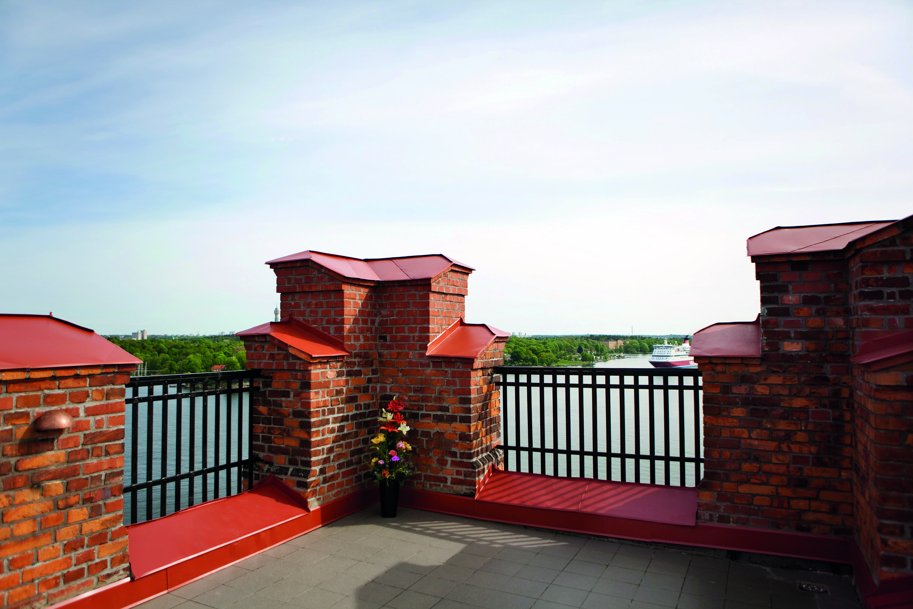 Rooftop terrace with red brick walls overlooking a scenic river view and distant ferry, featuring a potted plant with colorful flowers