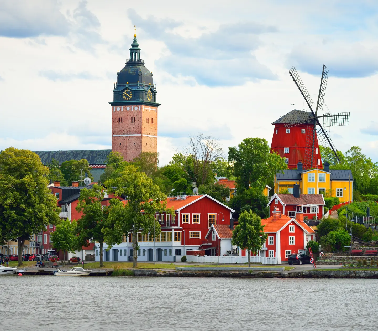 Vy över Strängnäs domkyrka och en traditionell röd väderkvarn i Strängnäs, med pittoreska röda hus längs vattnet.