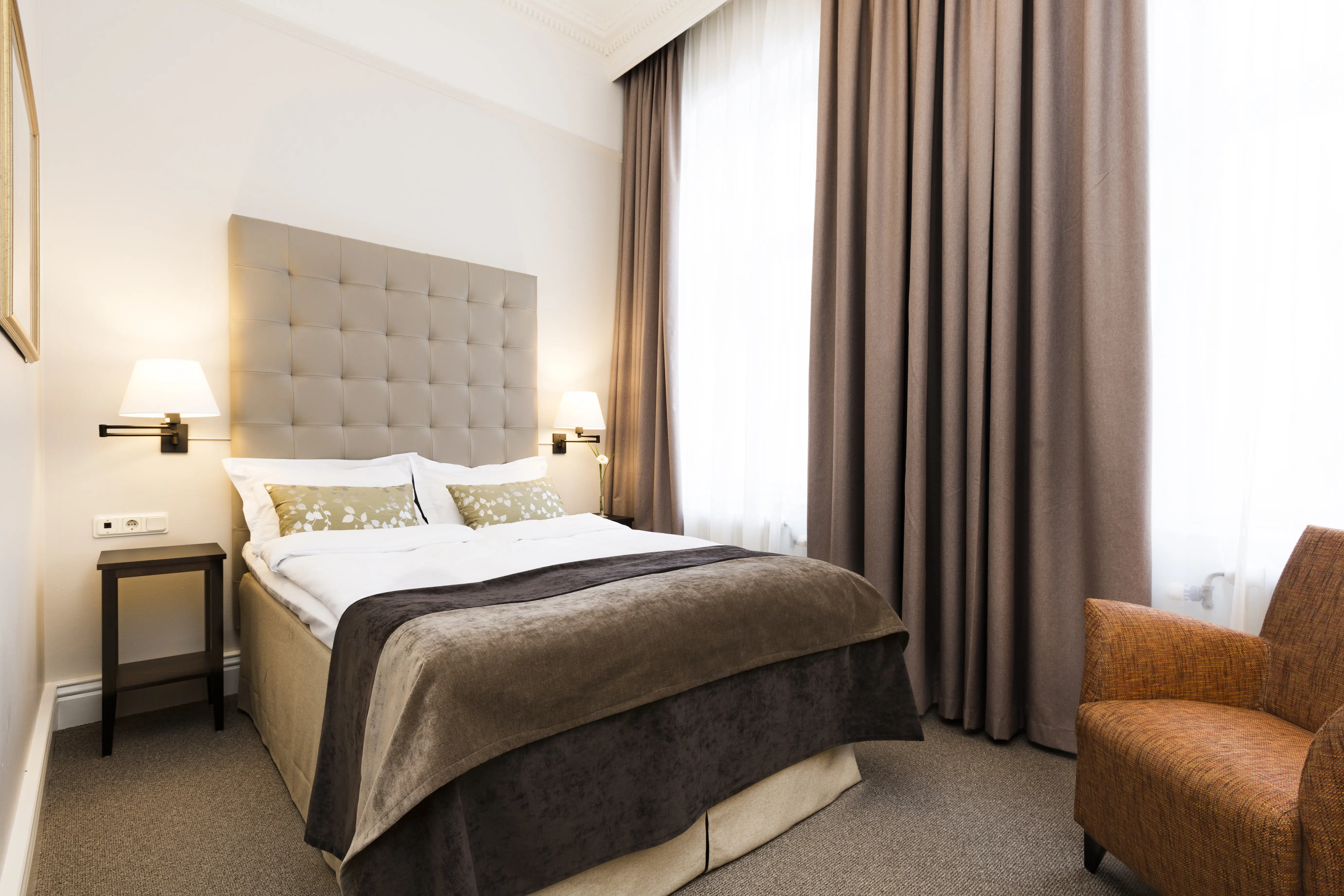 Ljust hotellrum med säng, grå sänggavel och fåtölj