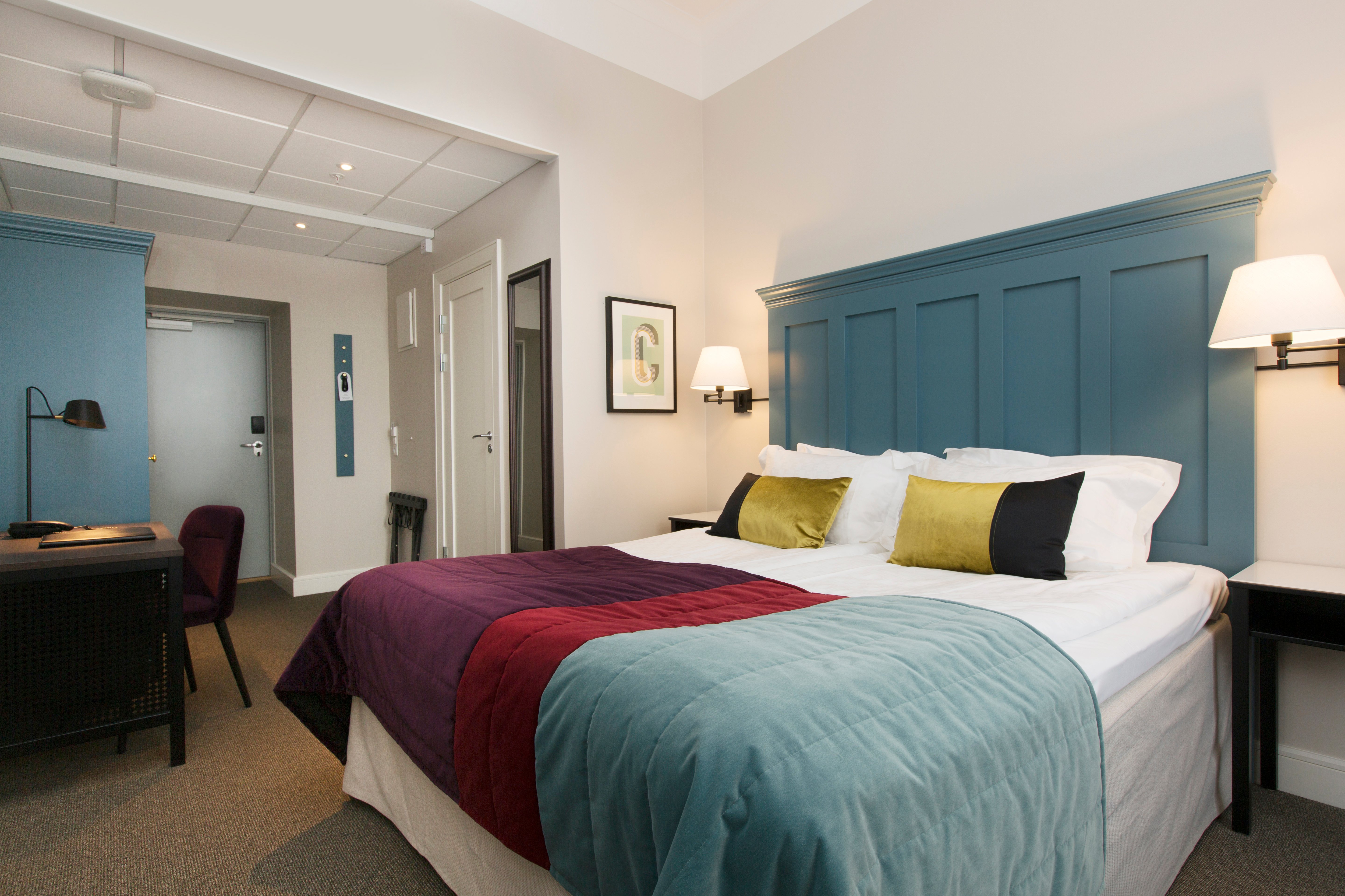 Bright hotel room with bed, large blue headboard and desk