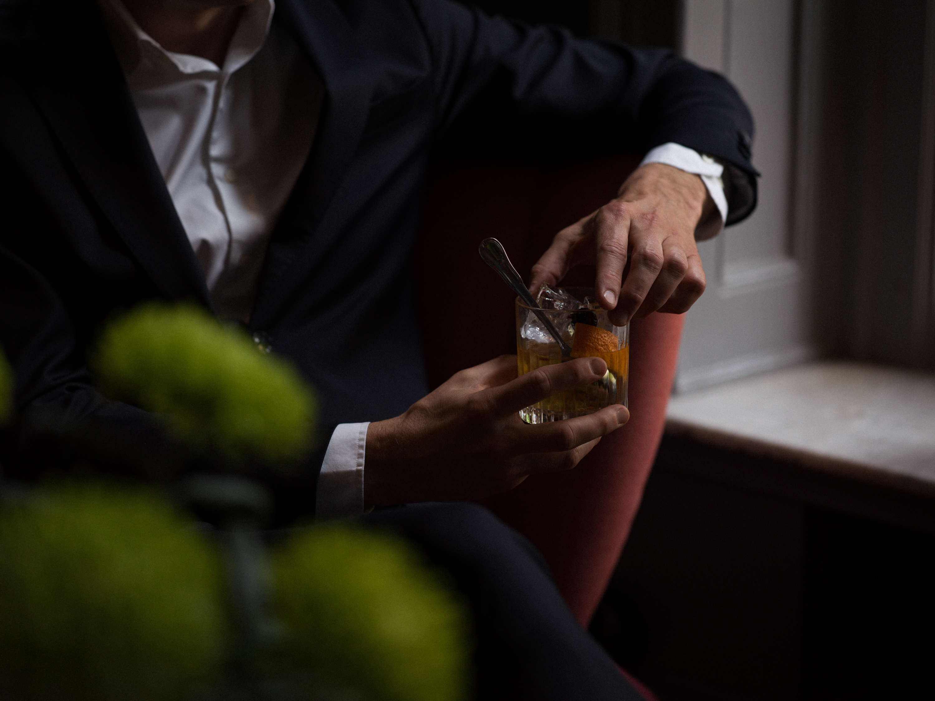 En person i kostym sitter ner och håller en cocktail i handen med en apelsinskalbit, i ett dämpat ljus