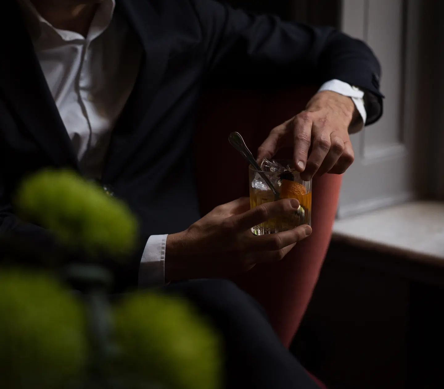 En person i kostym sitter ner och håller en cocktail i handen med en apelsinskalbit, i ett dämpat ljus