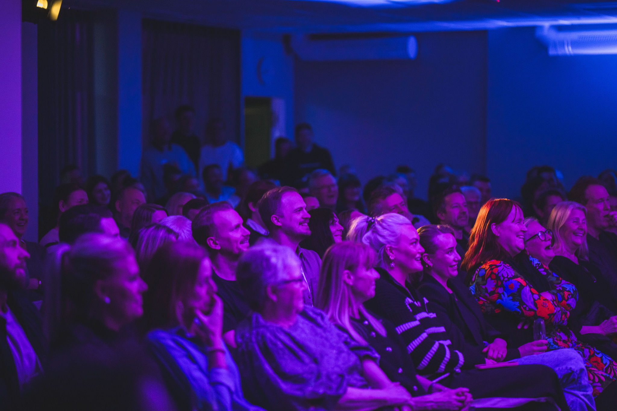 Publik i ett mörkt rum tittar på en scen, upplyst av blått och magenta ljus, ser engagerade och glada ut