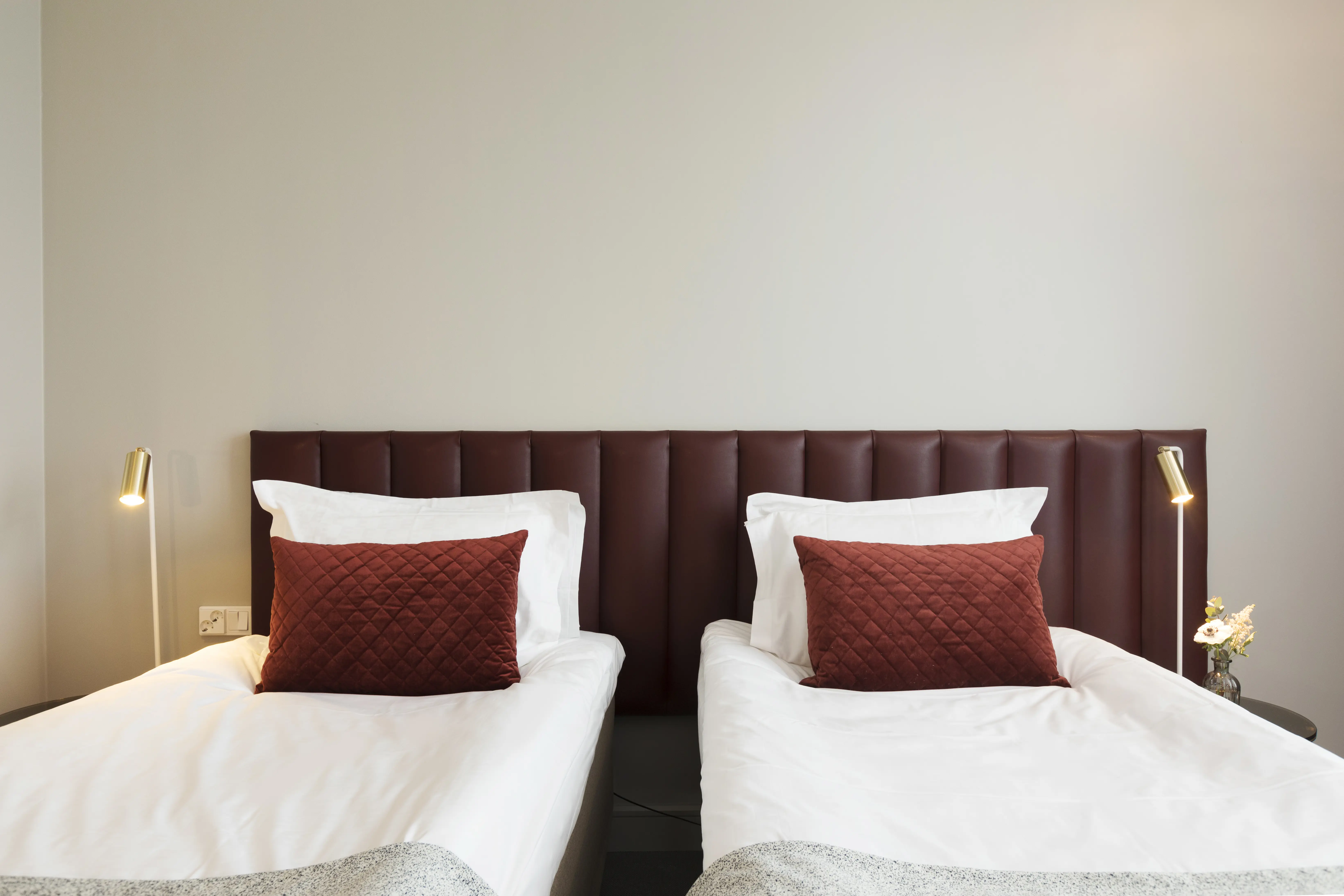 Ljust hotellrum med två enkelsängar bredvid varandra, vinröd sänggavel och små sänglampor