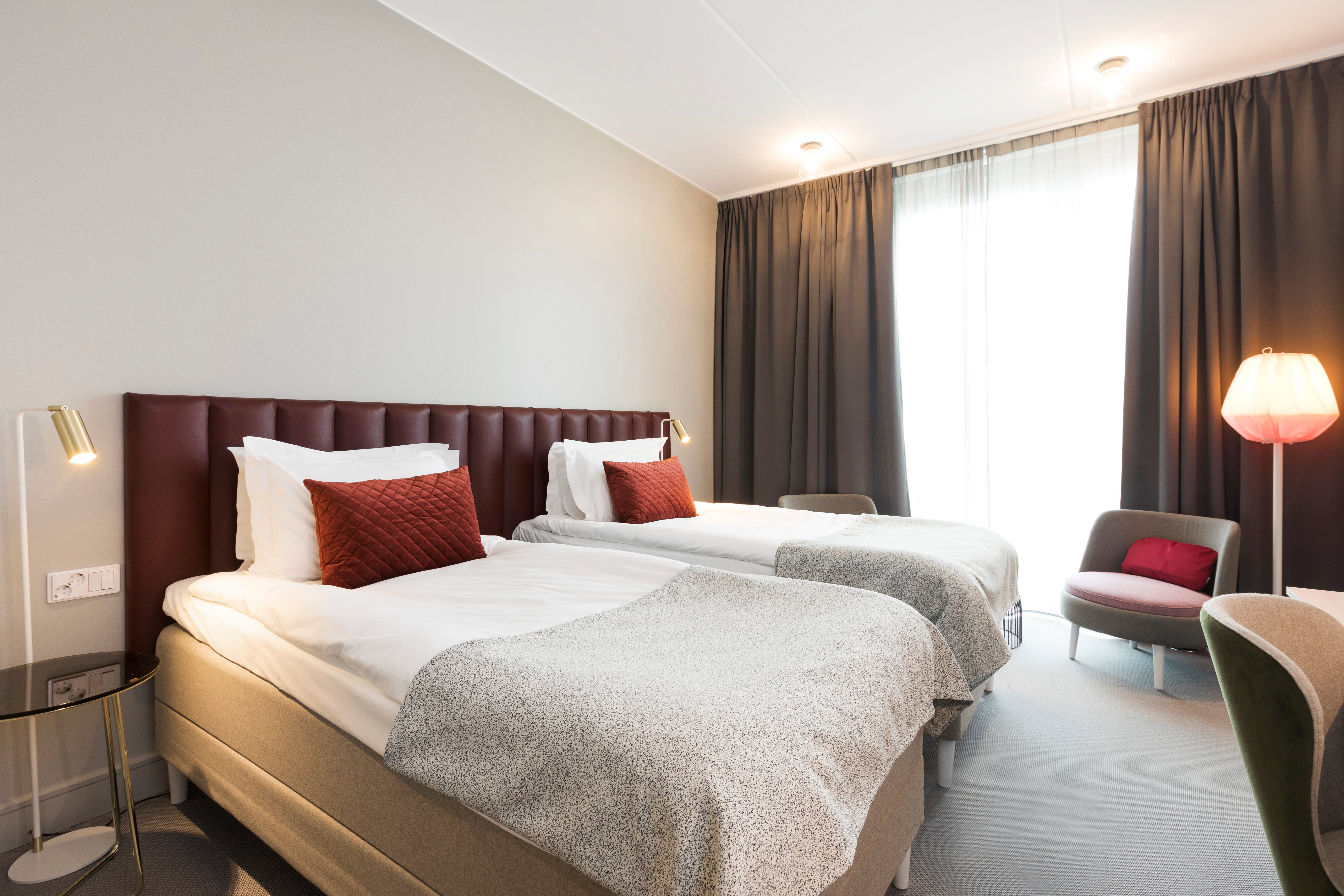 Bright hotel room with two single beds next to each other, burgundy headboard and armchairs