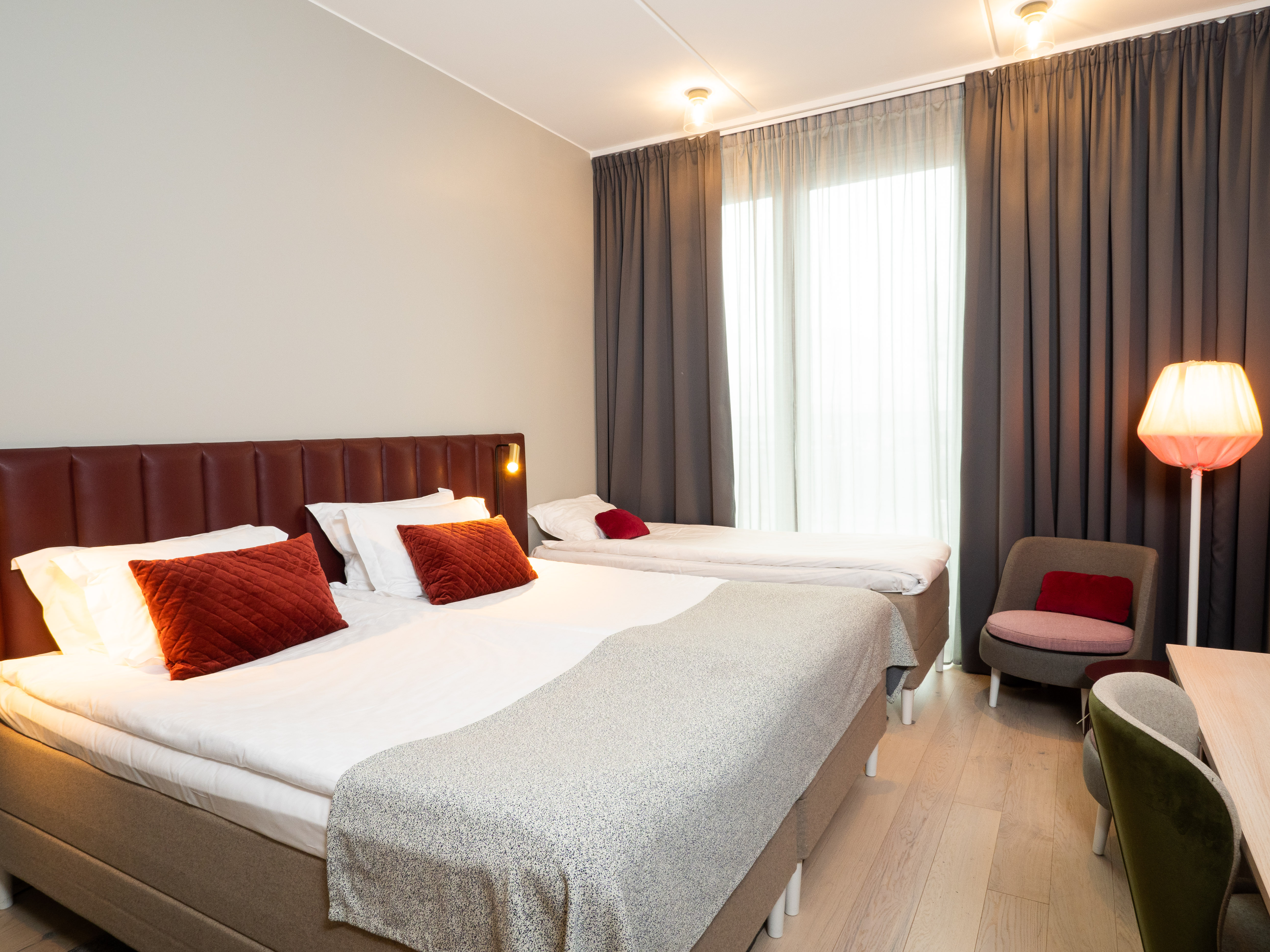 Hotellrum med en dubbelsäng, enkelsäng och modern inredning, inklusive en golvlampa, mjuka fåtöljer och mörkgråa gardiner.