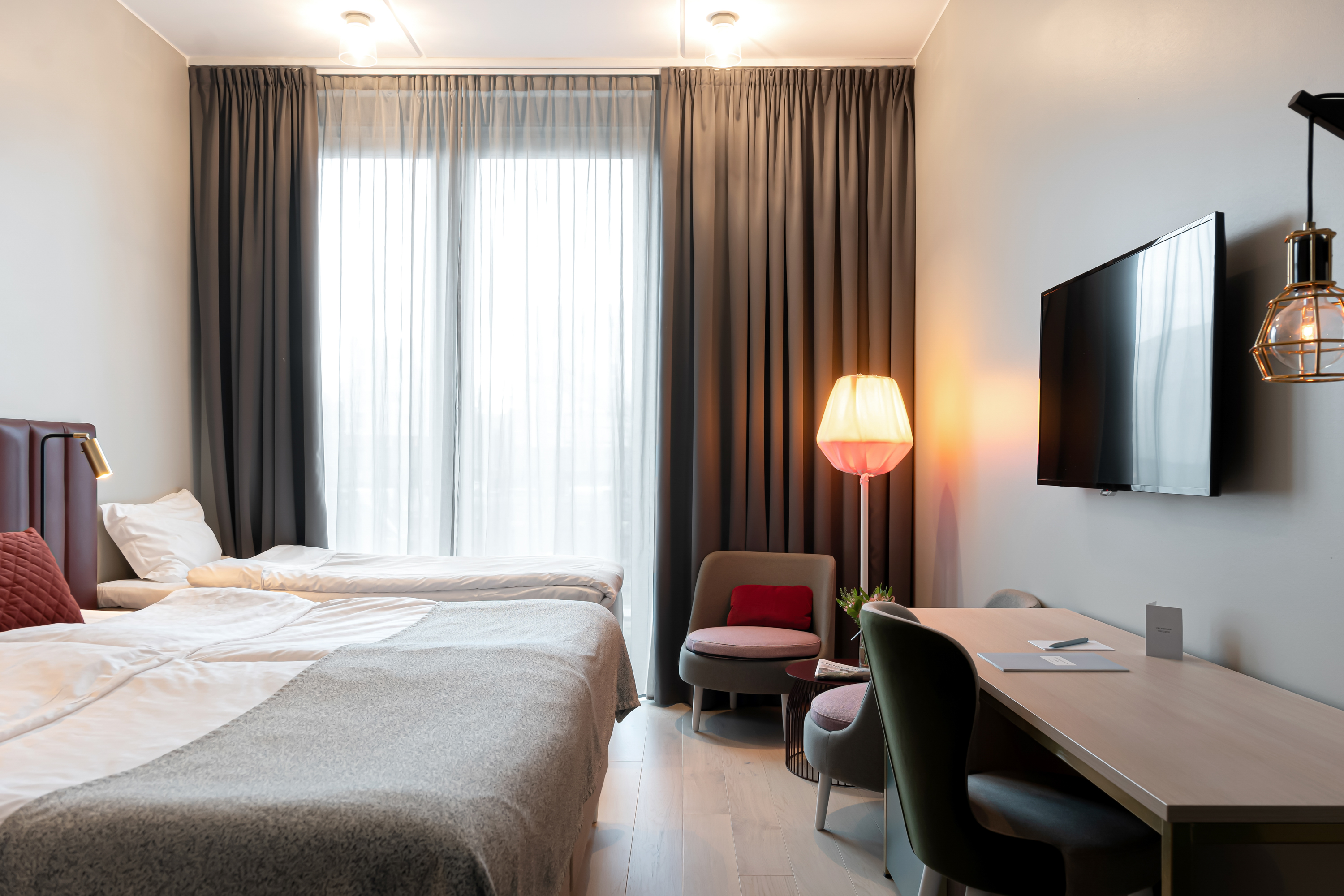 Hotellrum med en dubbelsäng, enkelsäng och modern inredning, inklusive en golvlampa, mjuka fåtöljer och mörkgråa gardiner.