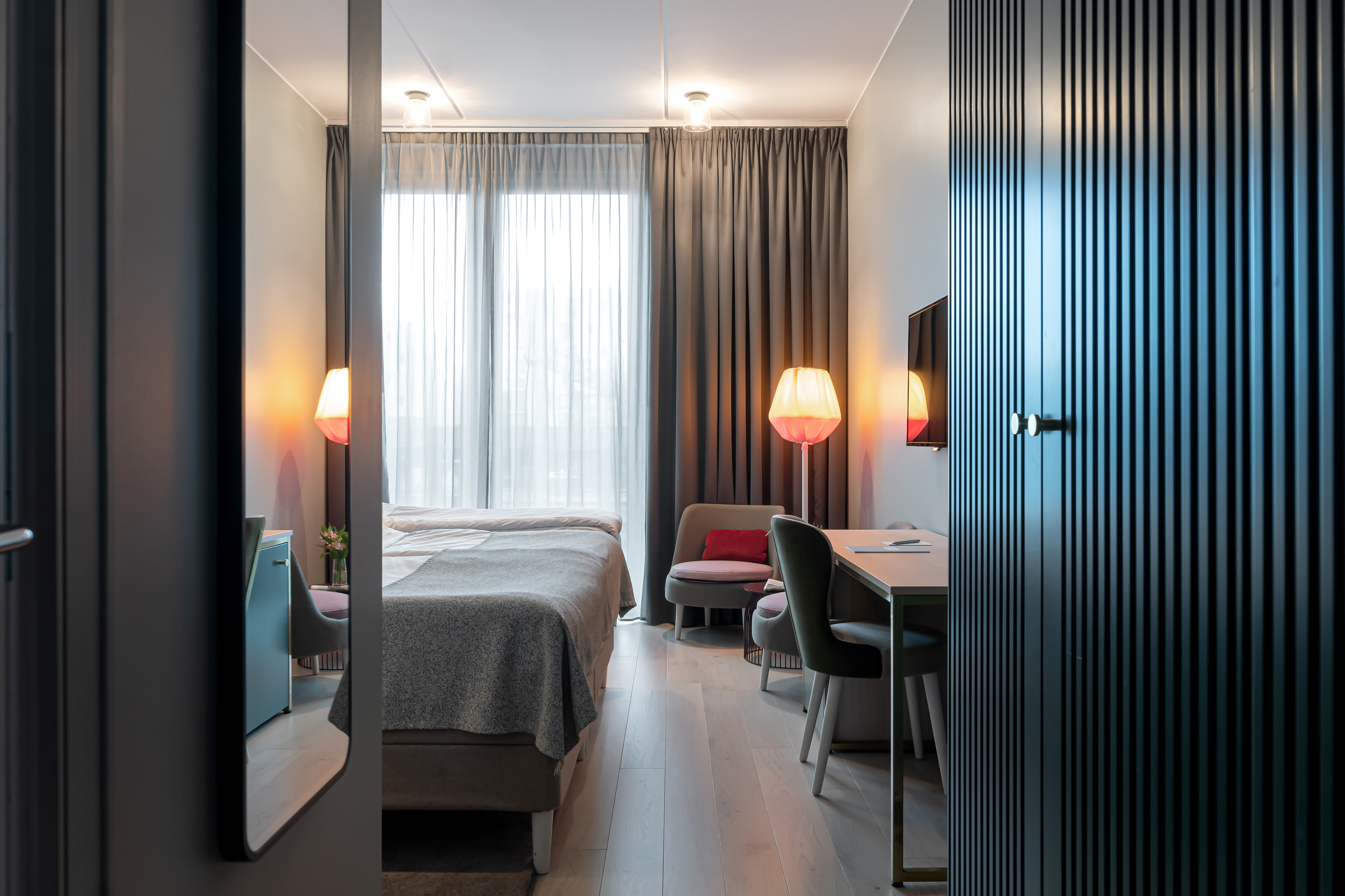 Modernt hotellrum med mysig belysning, dubbelsäng, skrivbord med stol och sittgrupp nära ett stort fönster med grå gardiner.