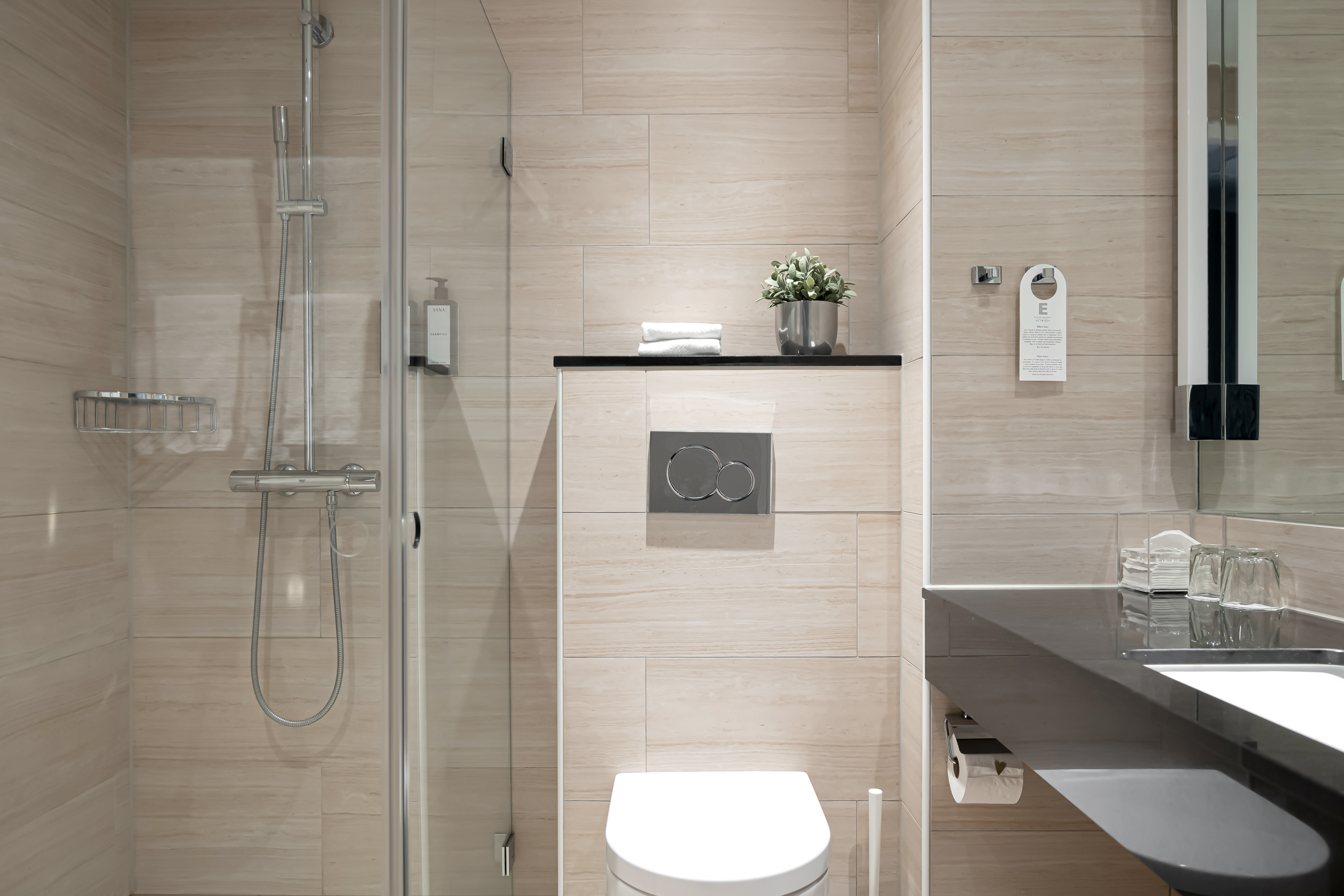 Modernt badrum med duschvägg i glas, toalett, svart handfat och ljusa kakelväggar. Dekorerat med en grön växt och en handduksstång.