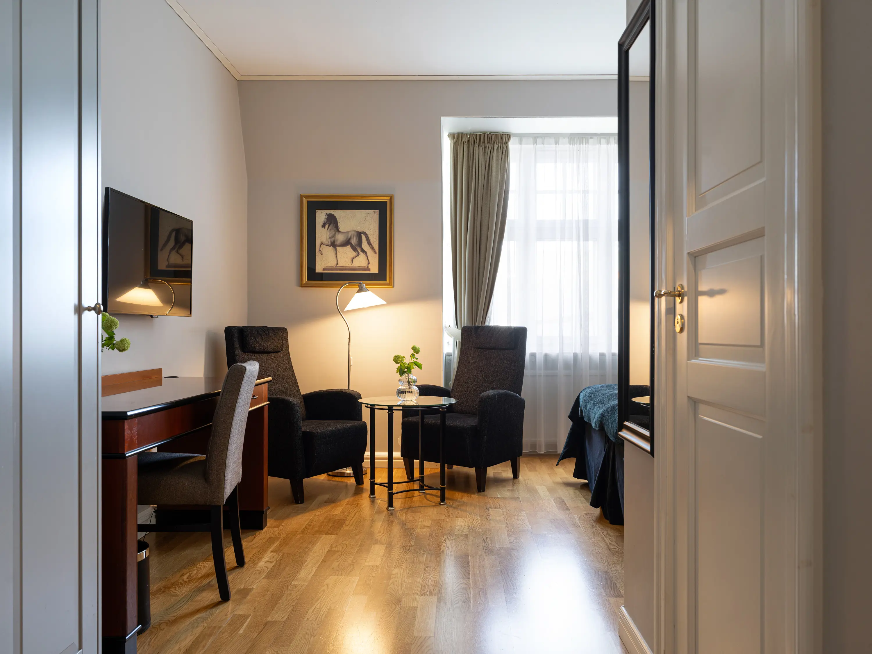 Elegant hotellrum med trägolv, två fåtöljer, skrivbord och platt-TV. Ljus fönsterbelysning med gardiner ger en sofistikerad känsla.