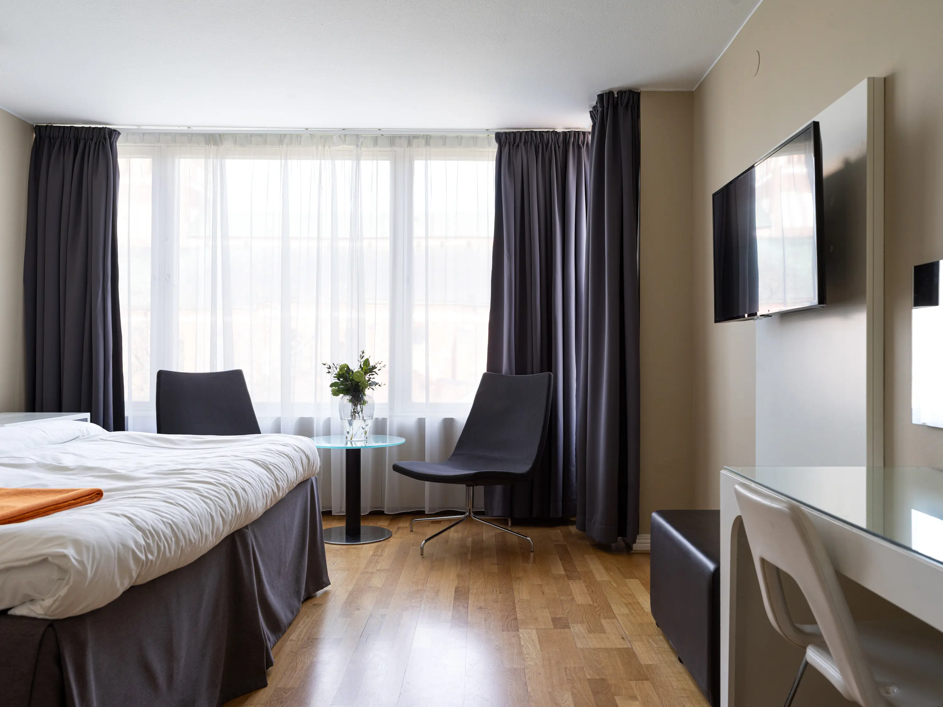 Modernt hotellrum med dubbelsäng, parkettgolv, två svarta fåtöljer och en väggmonterad TV.