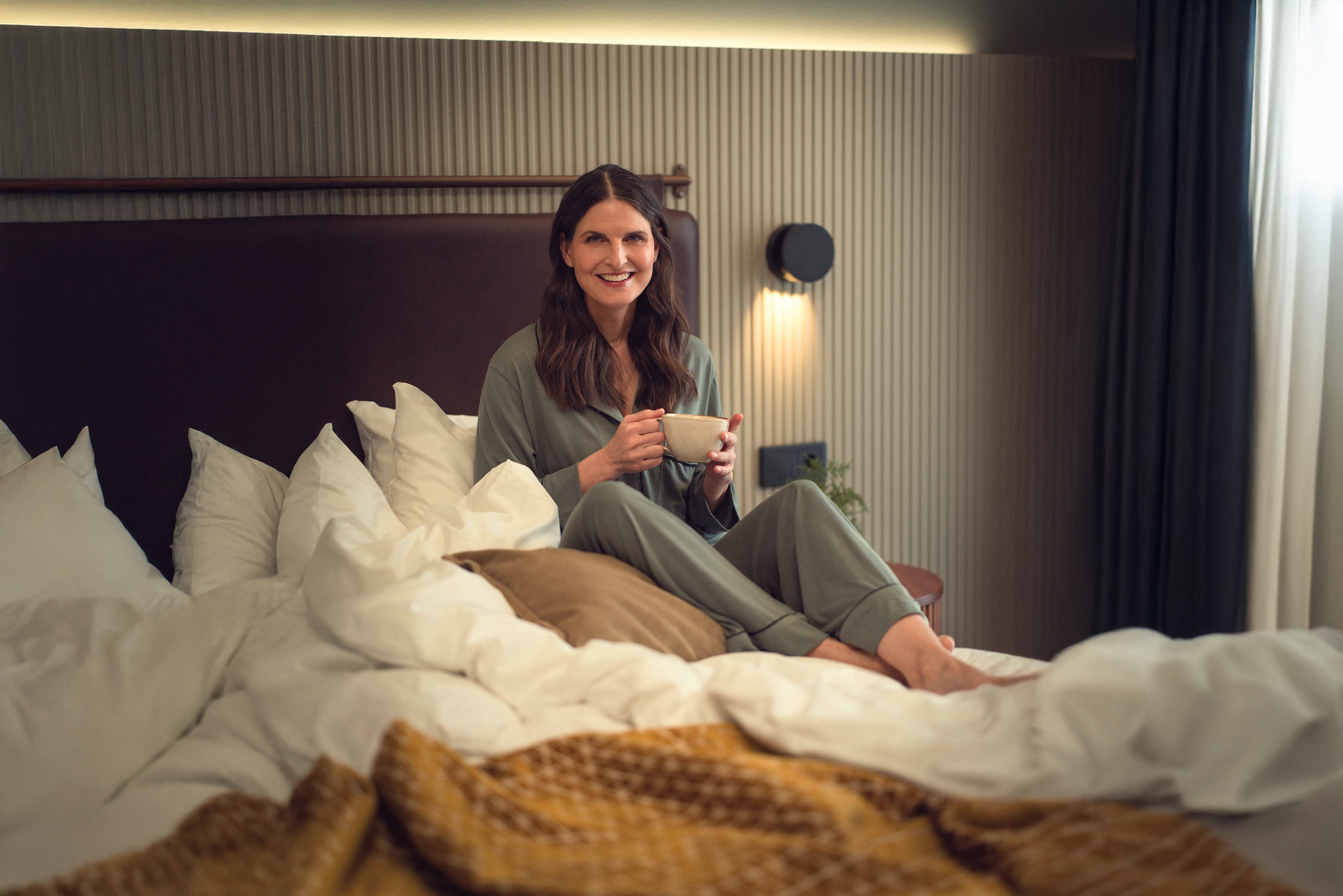 Woman in comfortable pajamas enjoying coffee on a cozy bed with soft pillows and warm lighting.