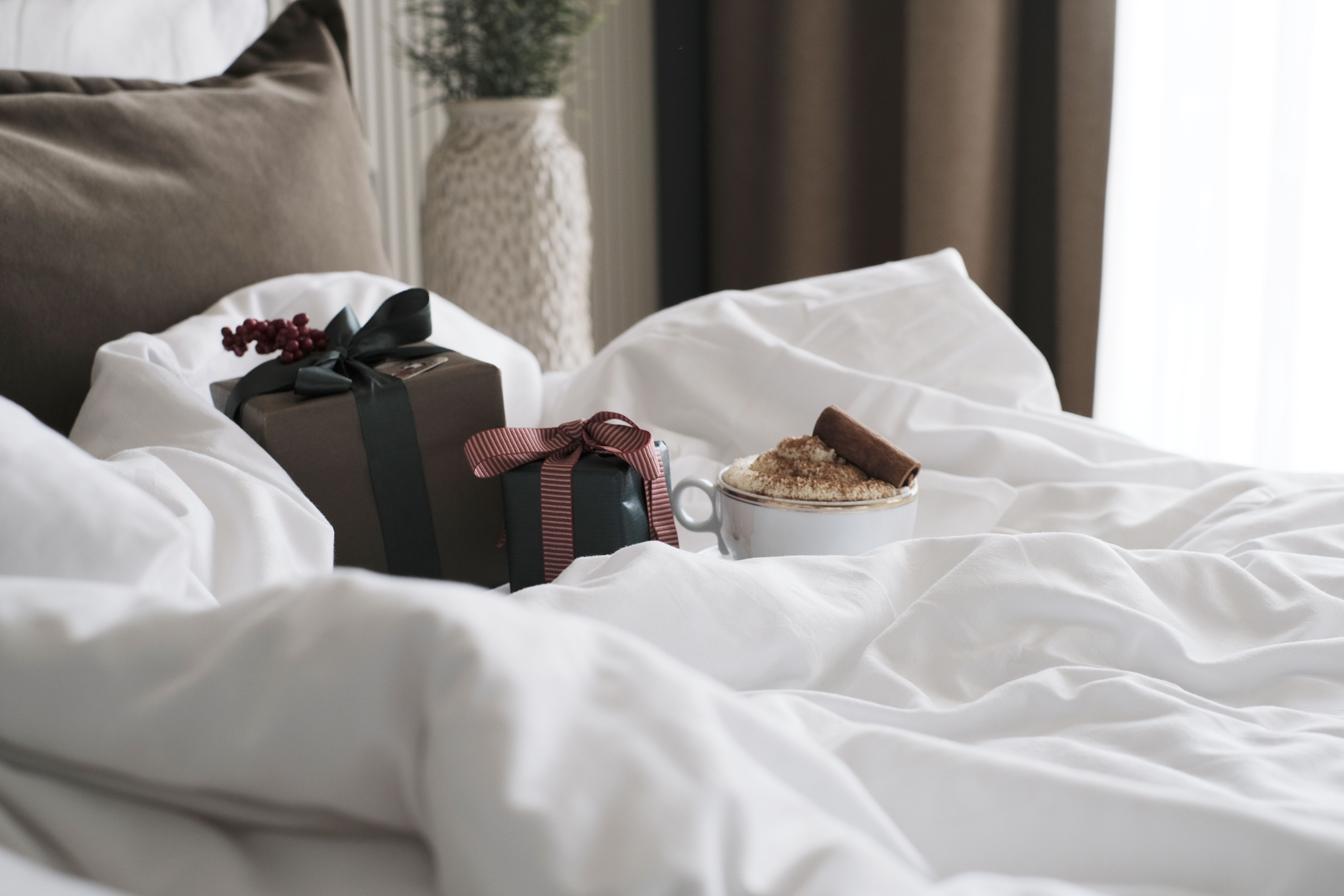 Julklapp med grönt band och röd present med randigt band på vit säng, bredvid en mugg cappuccino med kanelstång.