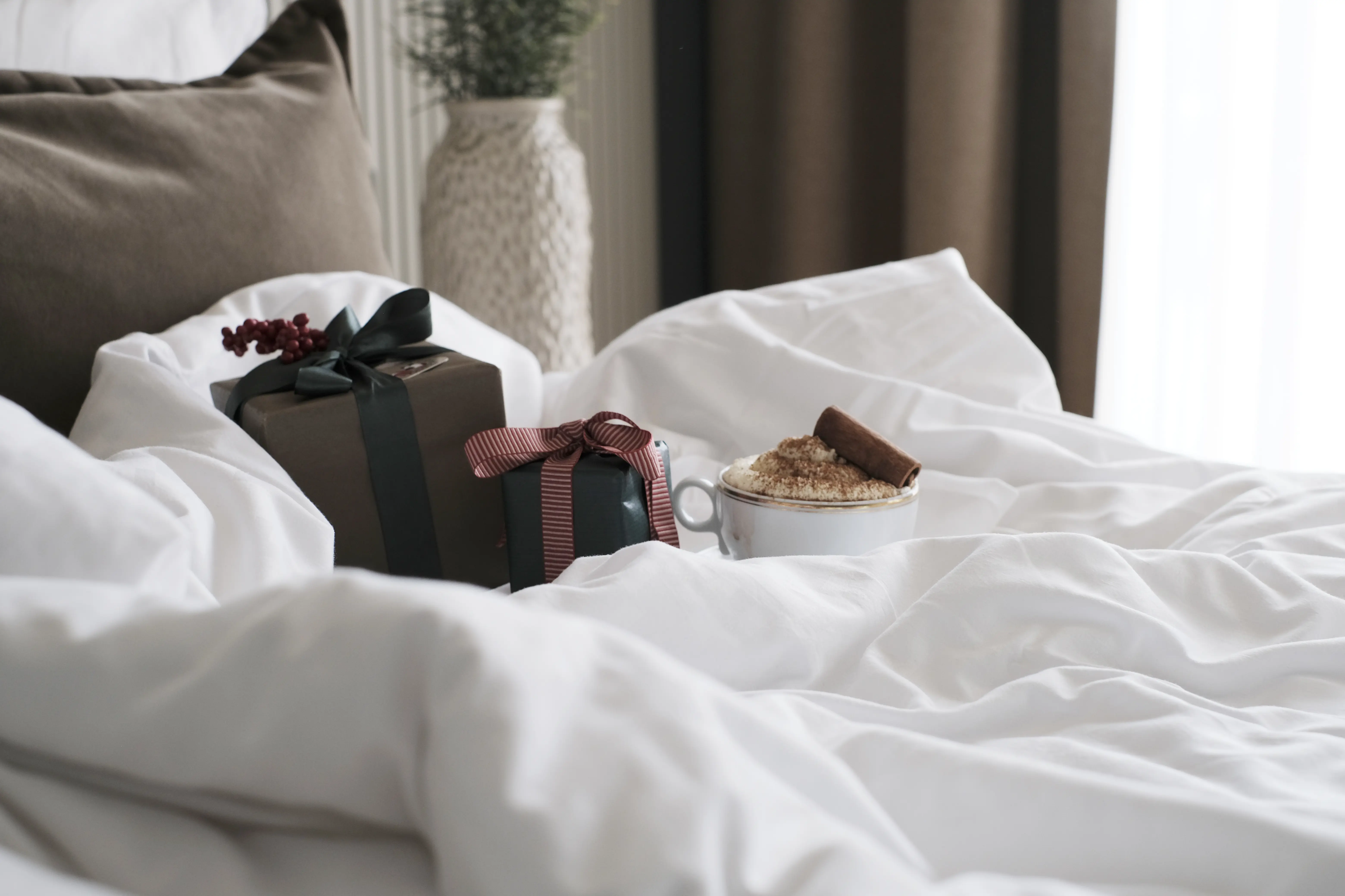 Cozy scene of a bed with white blankets, featuring a cup of coffee topped with cocoa and a cinnamon stick, alongside two gift boxes with bows, creating a warm, inviting atmosphere.