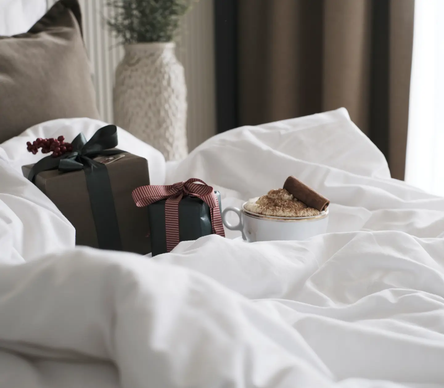 Julklapp med grönt band och röd present med randigt band på vit säng, bredvid en mugg cappuccino med kanelstång.