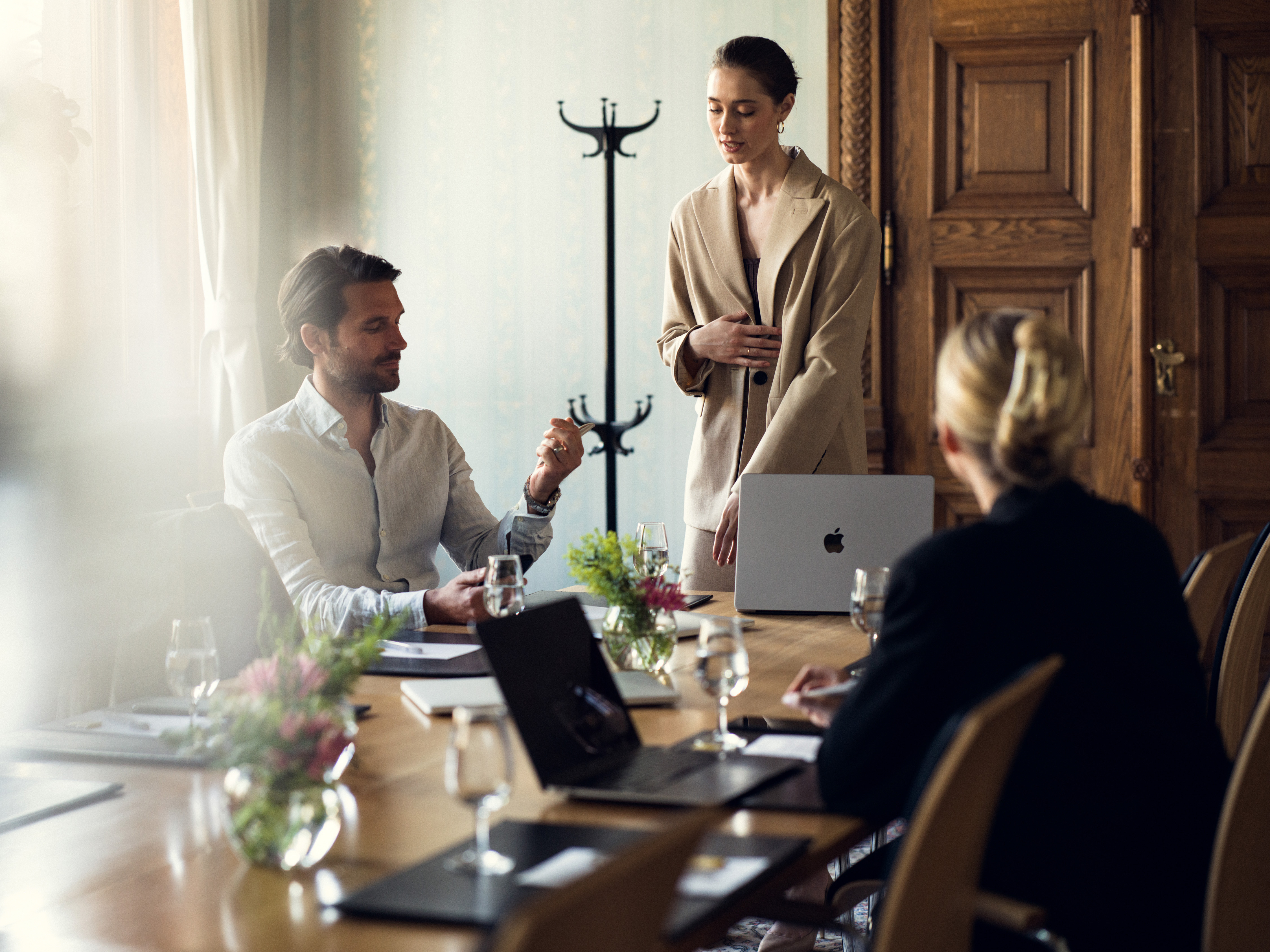 Business meeting in a conference room with three professionals, featuring laptops, documents, and floral arrangements on a wooden table.