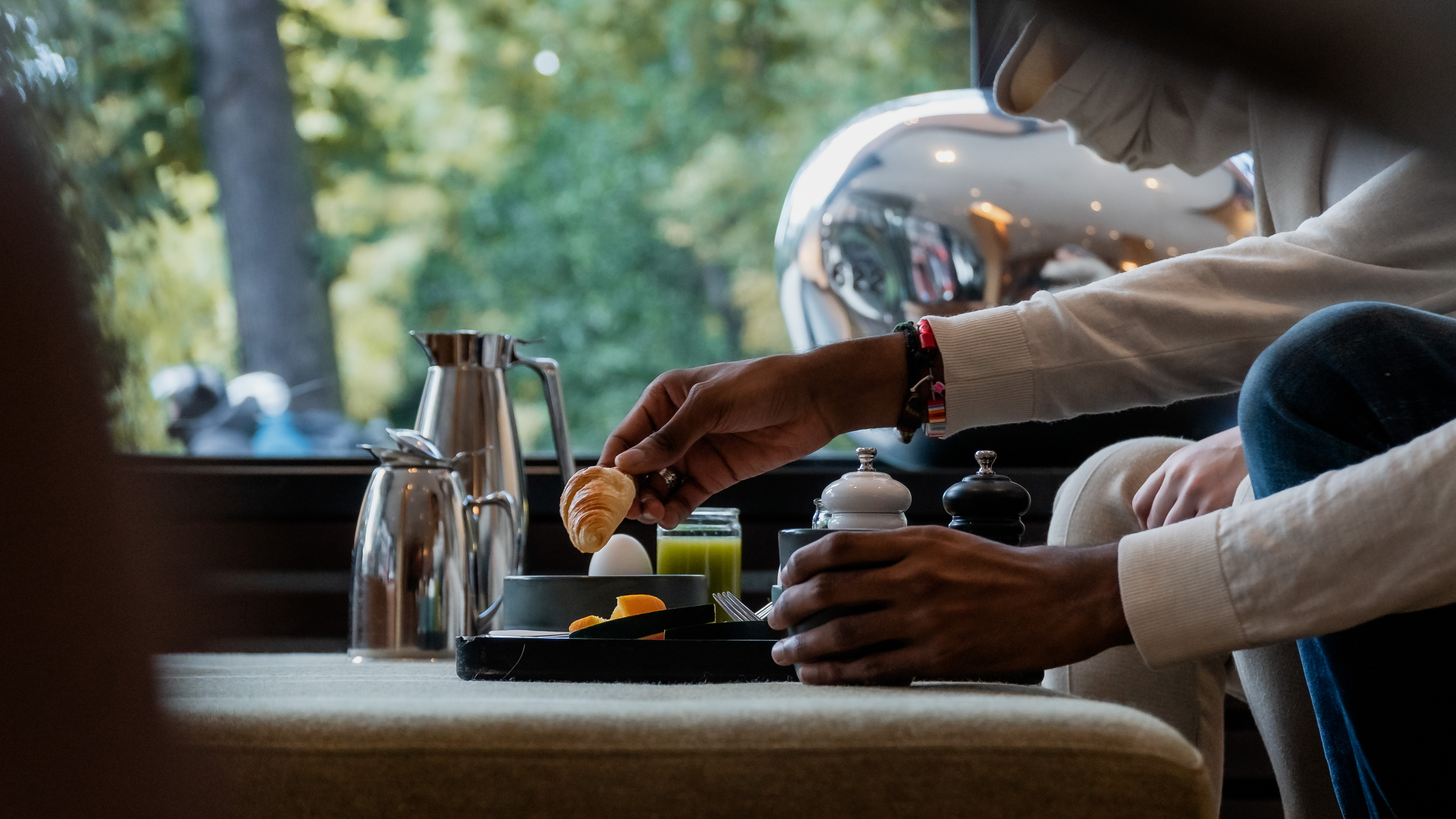 Person serverar frukost med kaffe, croissant och kokt ägg vid ett bord med naturligt ljus utifrån ett fönster