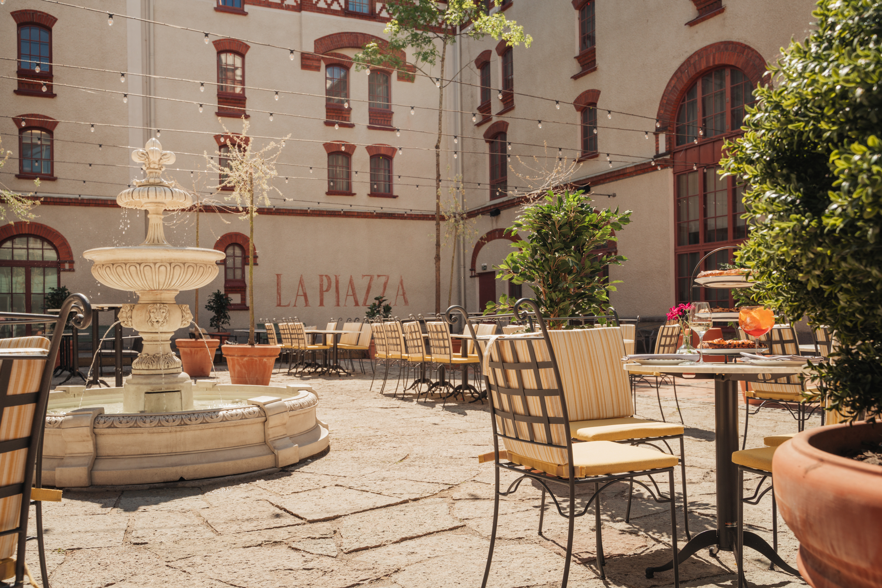 Fontän och uteservering på innergård med stolar och bord under solig himmel, "La Piazza" skrivet på väggen i bakgrunden