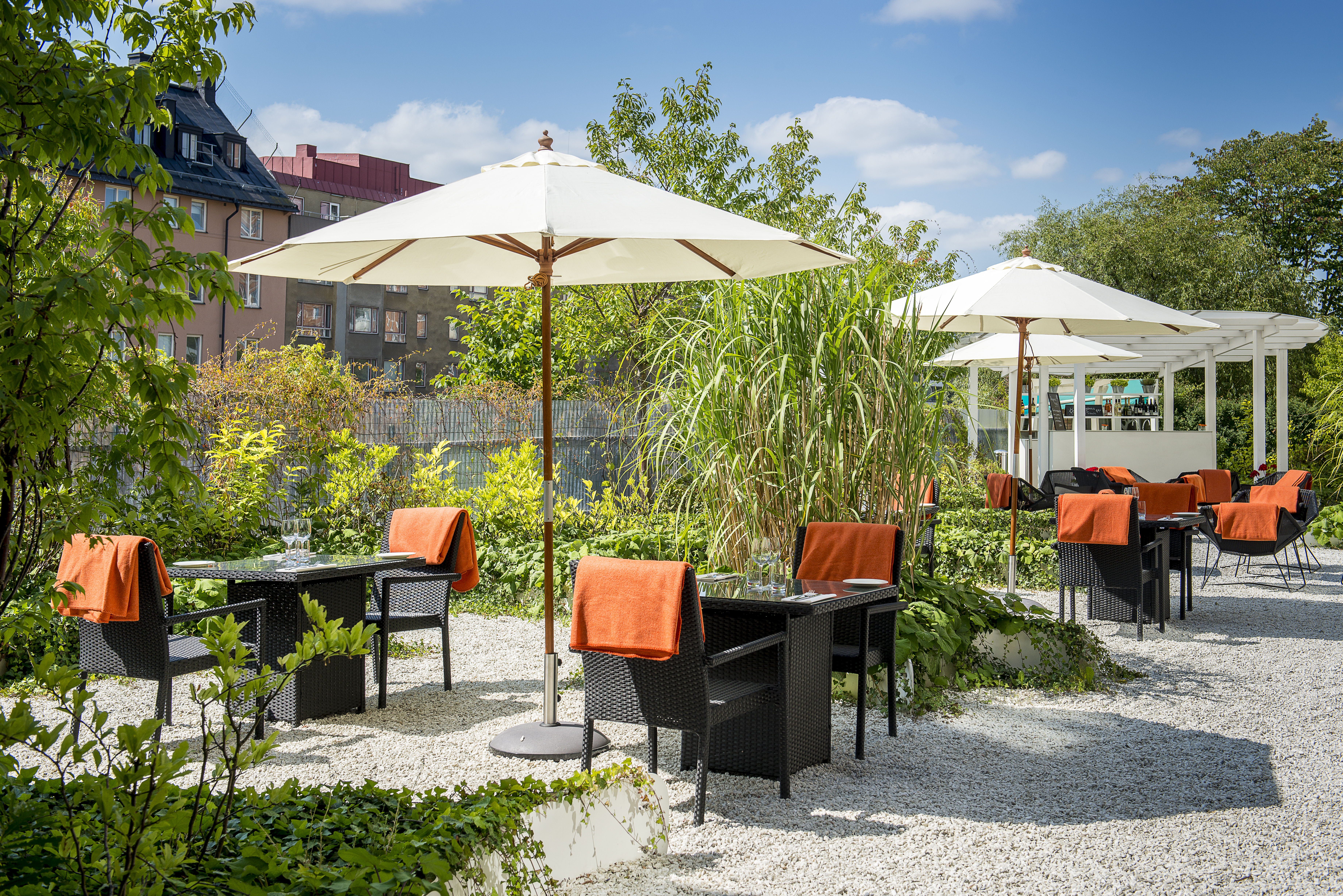 Uteservering i trädgårdsmiljö med parasoller och svarta caféstolar med orange tyg, omgiven av grönska och flerfamiljshus i bakgrunden under en klarblå himmel