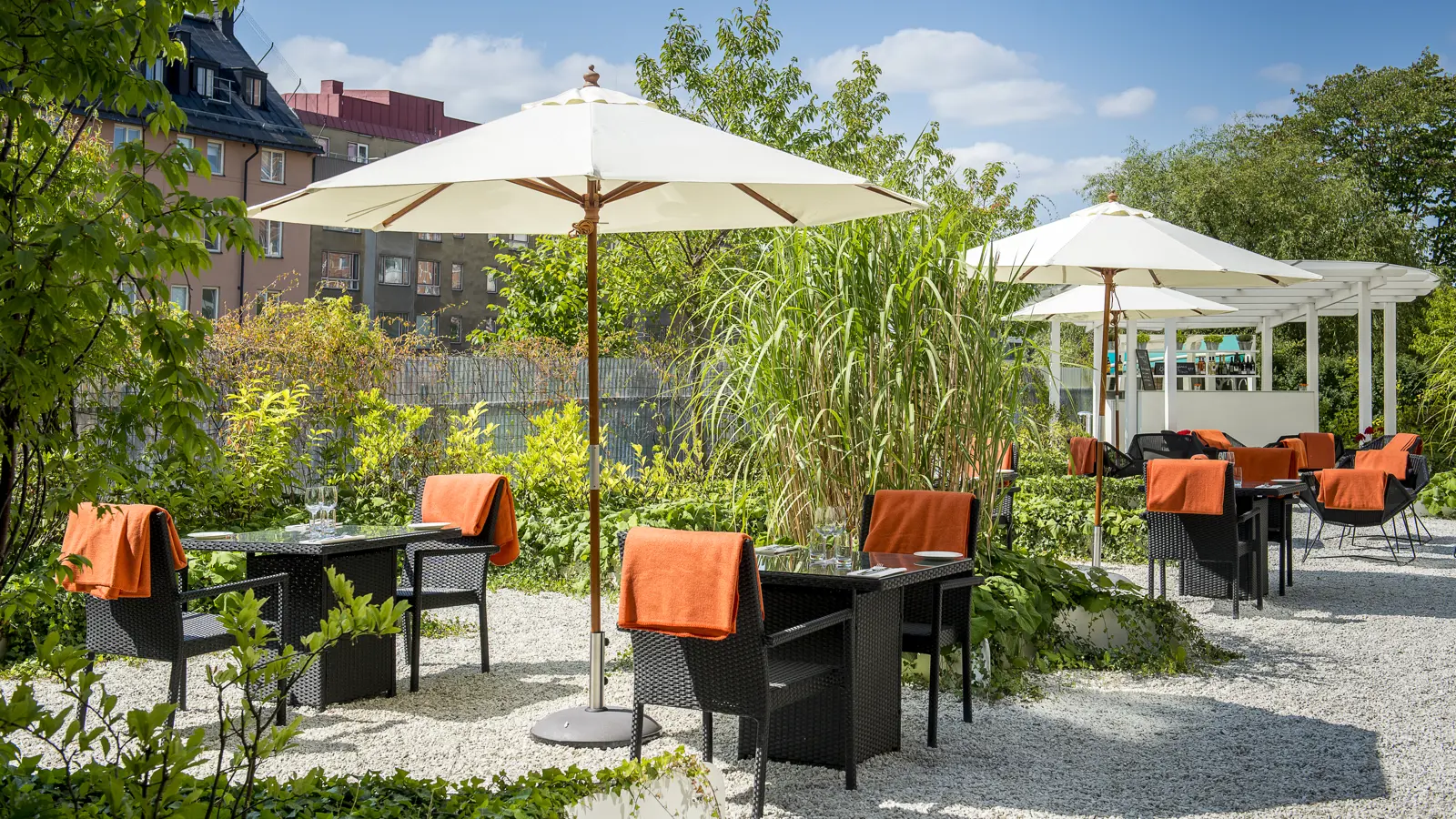 Uteservering i trädgårdsmiljö med parasoller och svarta caféstolar med orange tyg, omgiven av grönska och flerfamiljshus i bakgrunden under en klarblå himmel