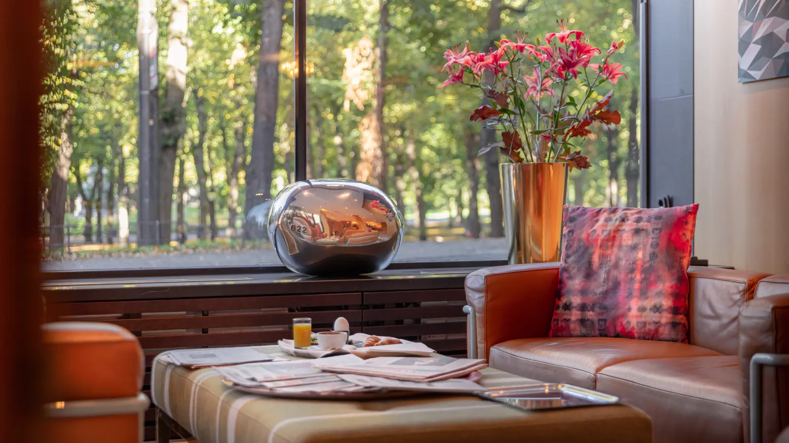 Modernt vardagsrum med lädermöbler och färgglada kuddar, stora fönster med utsikt över grönska, samt ett soffbord med tidningar och drycker