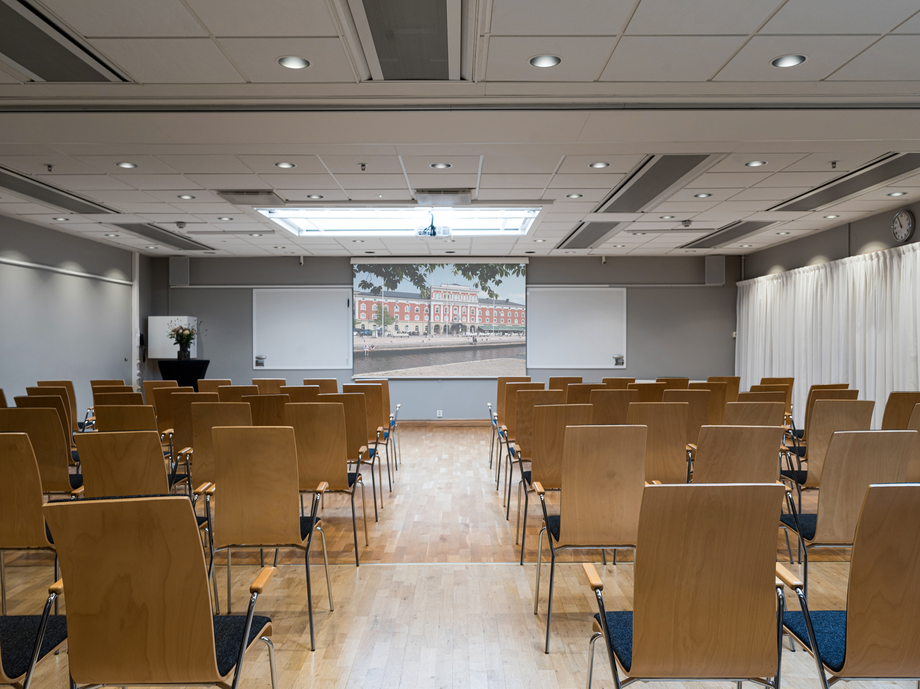 Conference room setup with wooden chairs facing a screen and whiteboards, featuring a modern ceiling design and natural lighting.