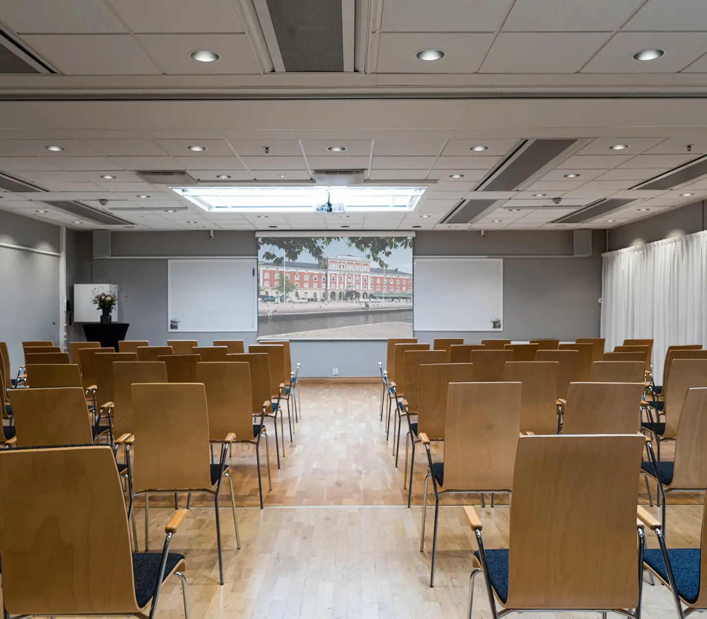 Conference room setup with wooden chairs facing a screen and whiteboards, featuring a modern ceiling design and natural lighting.