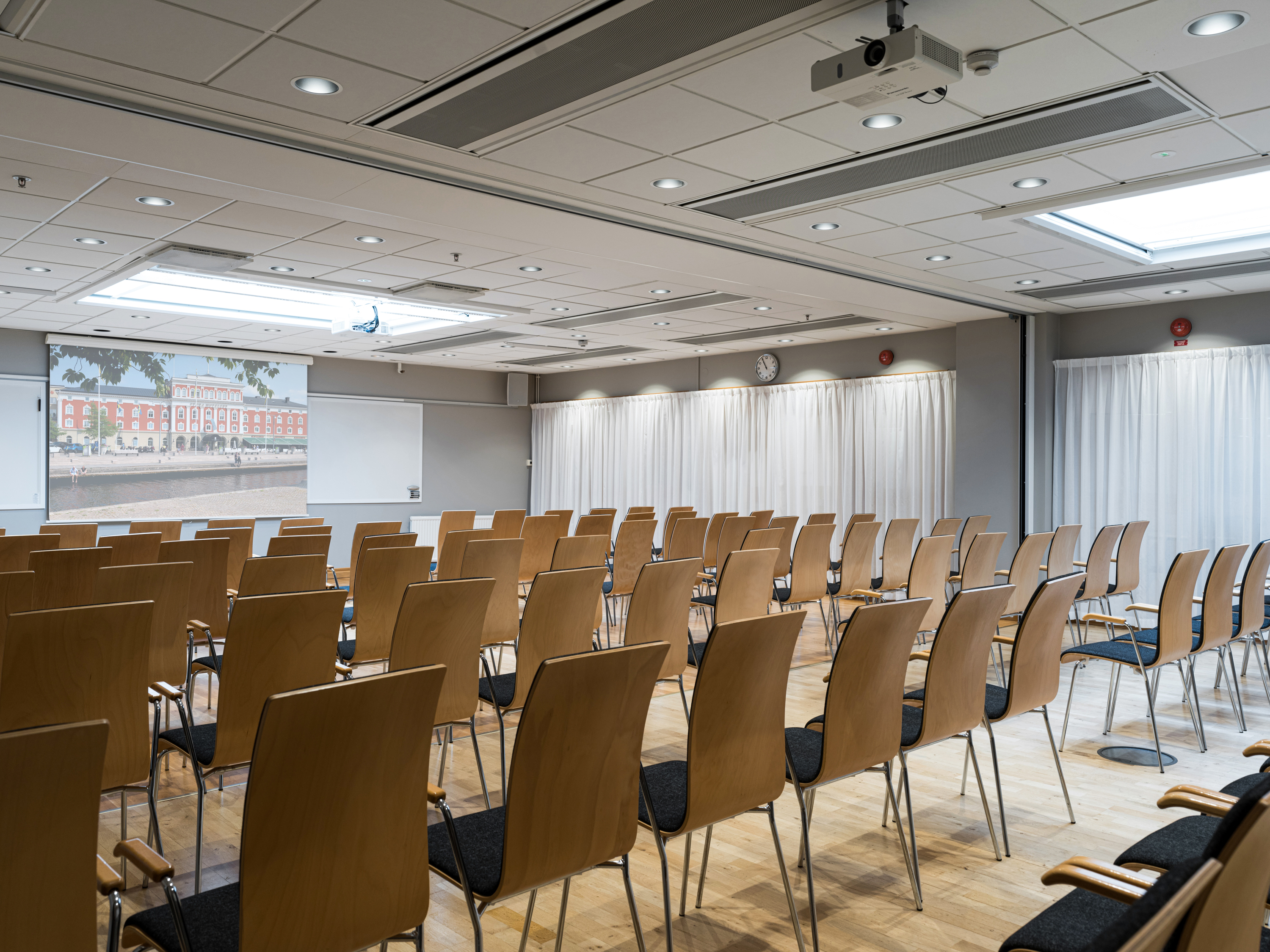 Modern conference room with wooden chairs arranged in rows, projection screen at the front, and bright overhead lighting.