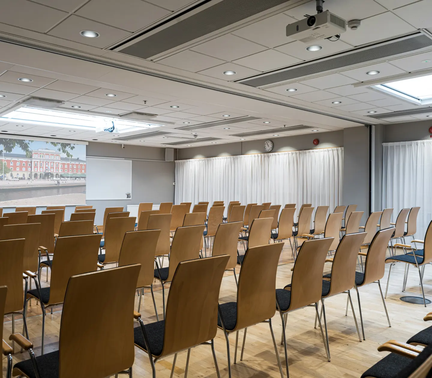 Modern conference room with wooden chairs arranged in rows, projection screen at the front, and bright overhead lighting.