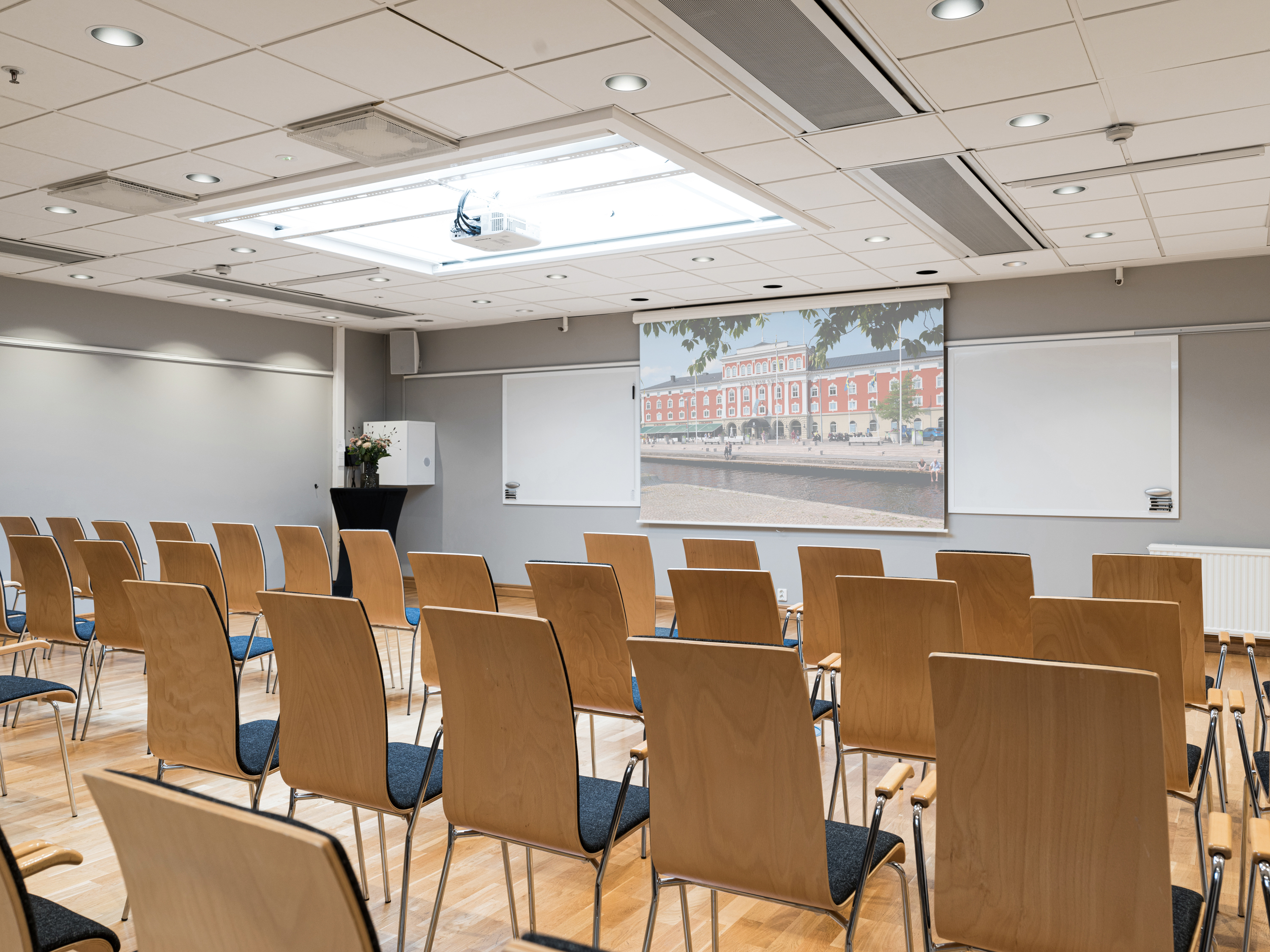 Modern conference room with rows of empty wooden chairs facing a large presentation screen and whiteboard, ideal for business meetings and seminars.