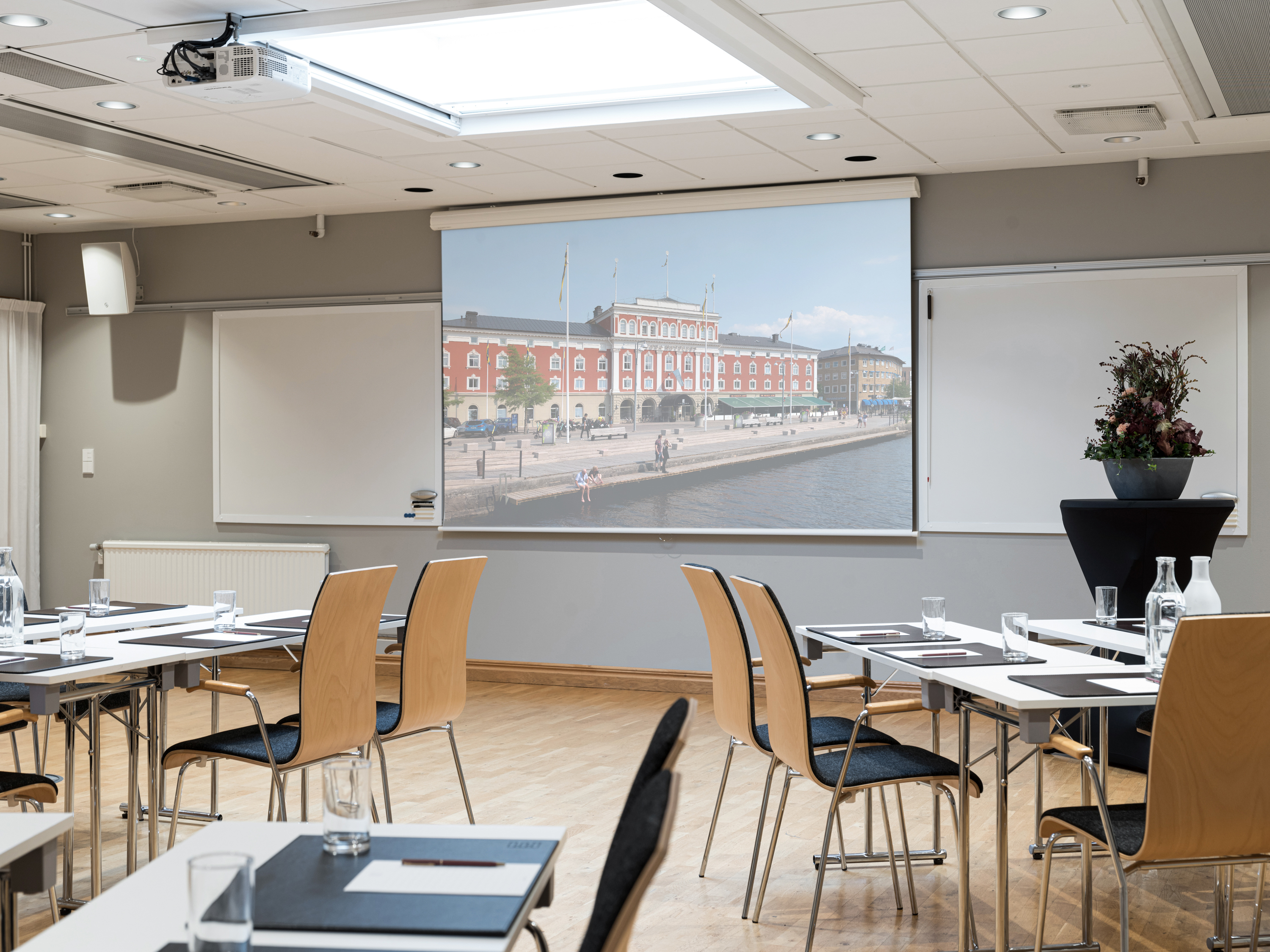 Conference room set up for a meeting with chairs, tables, and water glasses. A large projector screen displays an image of a historic red building by a waterfront.
