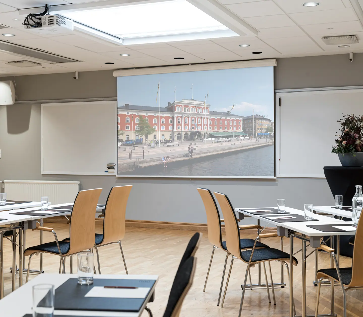 Conference room set up for a meeting with chairs, tables, and water glasses. A large projector screen displays an image of a historic red building by a waterfront.