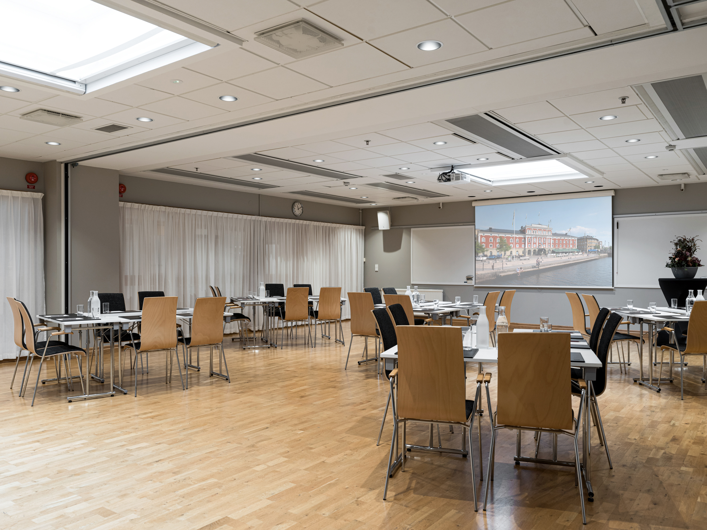 Modern conference room with wooden flooring, arranged tables, and chairs. A large screen displays an urban waterfront image.