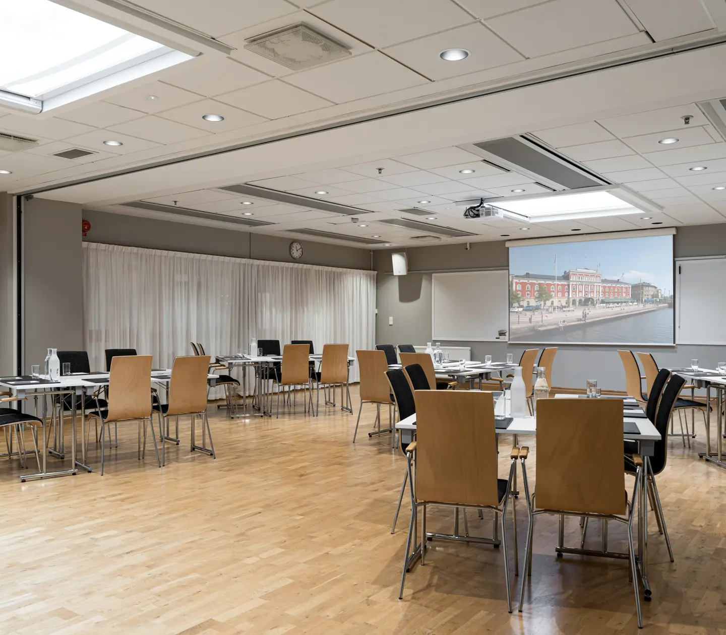 Modern conference room with wooden flooring, arranged tables, and chairs. A large screen displays an urban waterfront image.