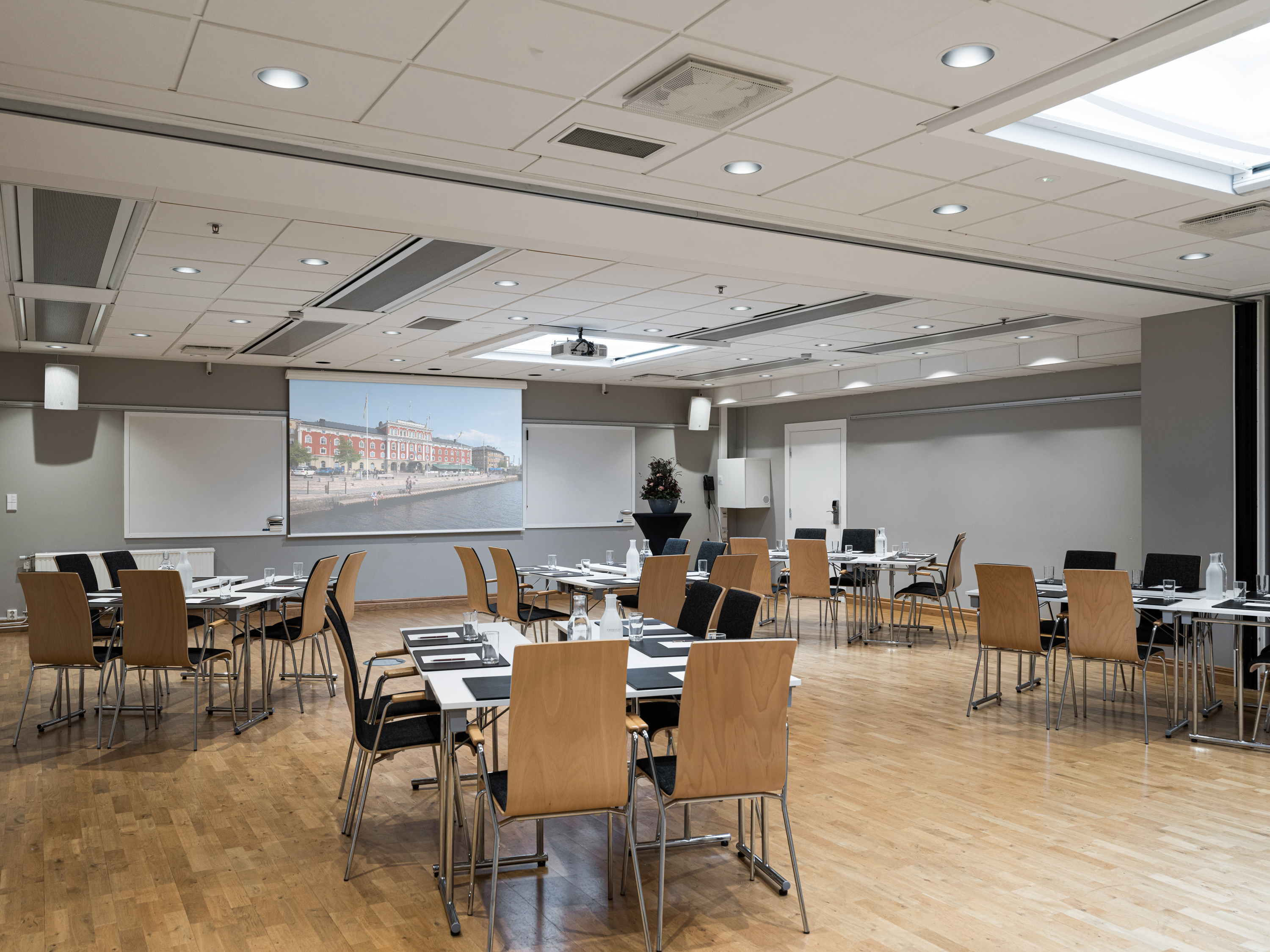 Modern conference room with wooden flooring, neatly arranged tables and chairs, a projector screen, and bright lighting, ideal for meetings and presentations.