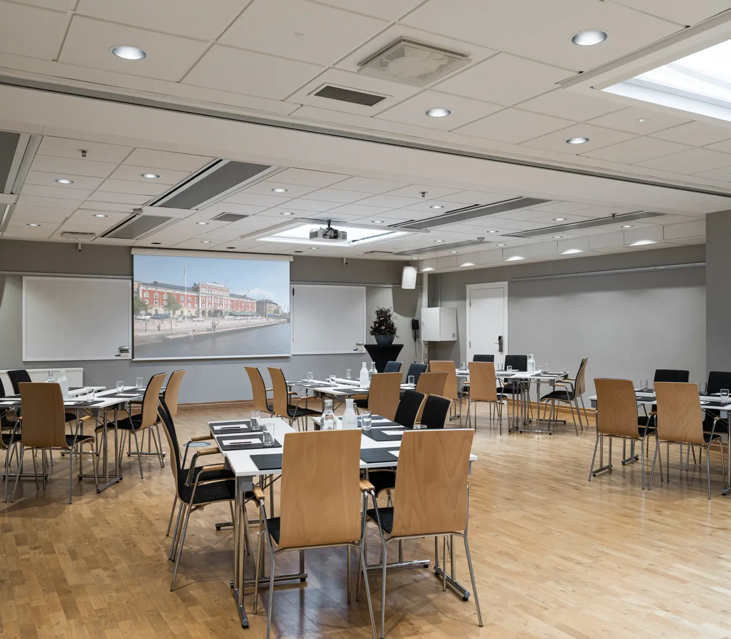 Modern conference room with wooden flooring, neatly arranged tables and chairs, a projector screen, and bright lighting, ideal for meetings and presentations.