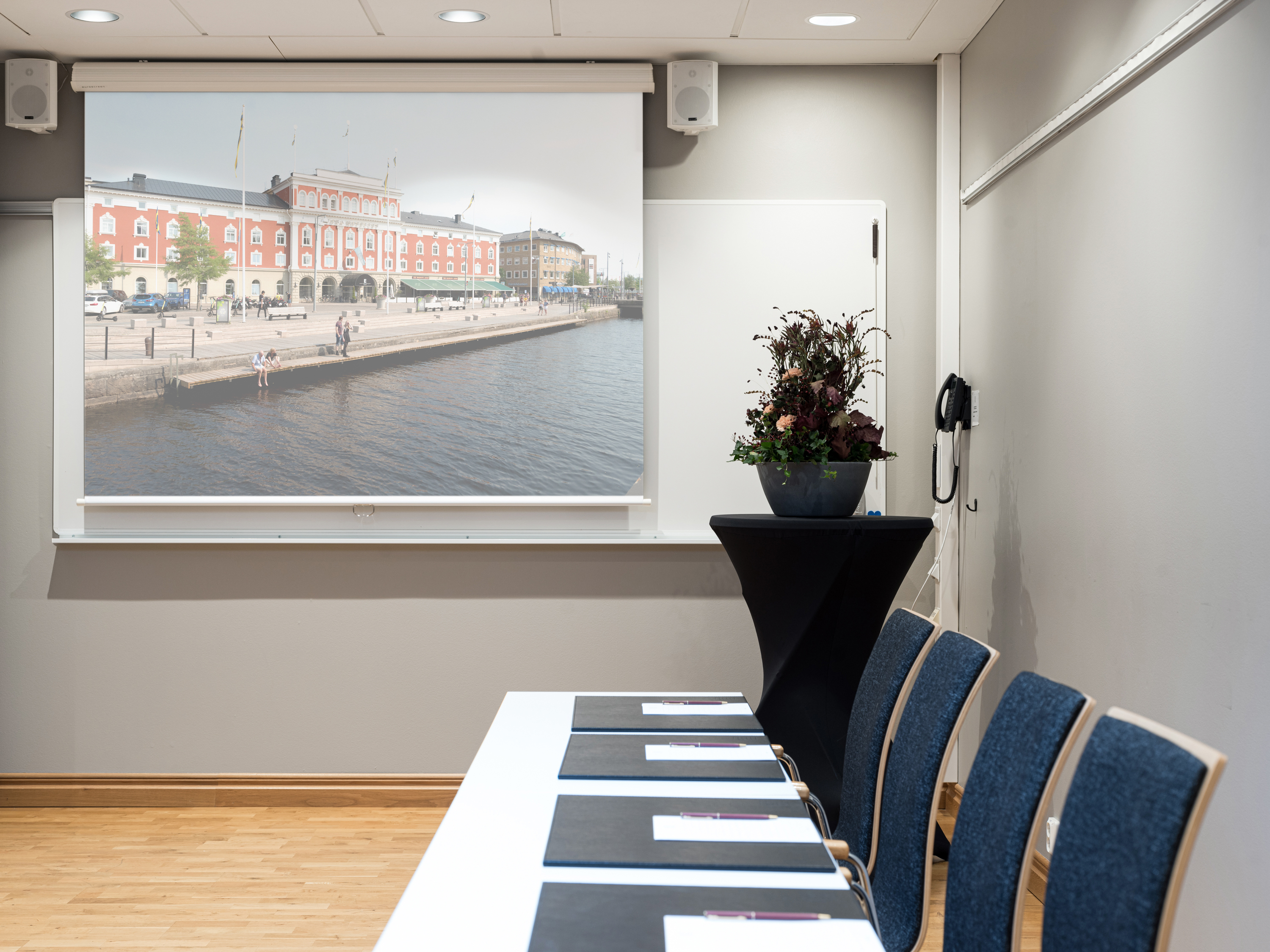 Conference room interior with a projection screen displaying a city waterfront scene, cushioned chairs, a long table with notepads, and a decorative plant.