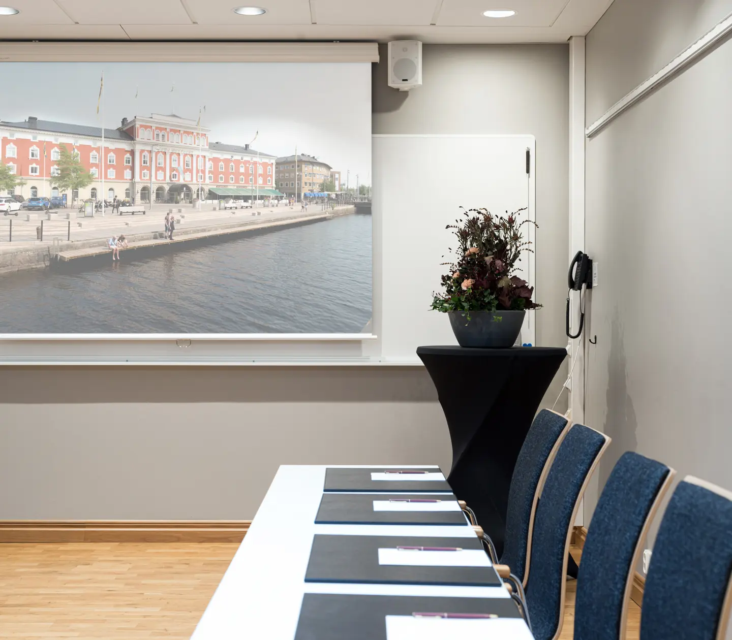 Conference room interior with a projection screen displaying a city waterfront scene, cushioned chairs, a long table with notepads, and a decorative plant.
