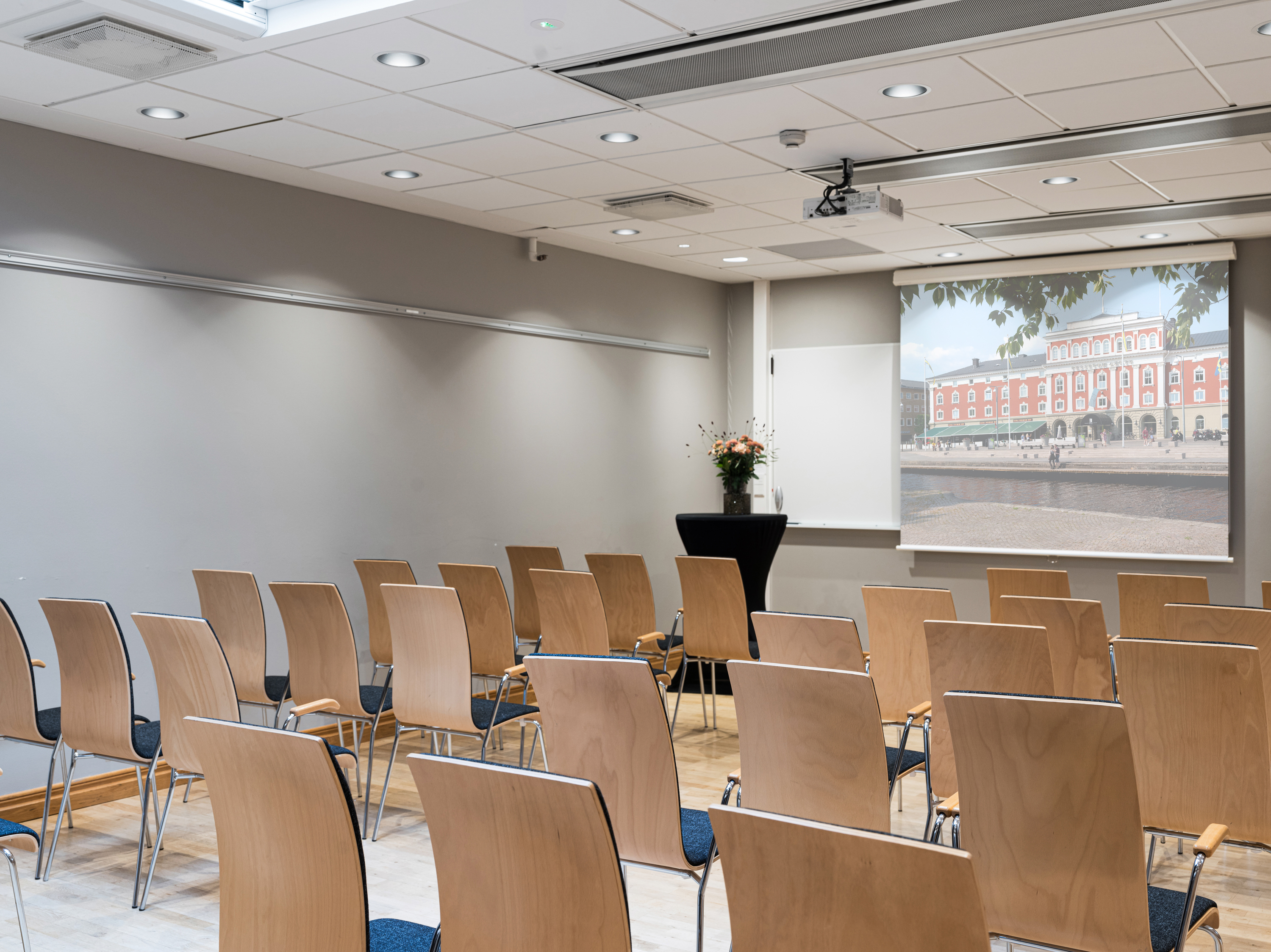 Modern conference room setup with rows of wooden chairs facing a projector screen displaying an exterior view of a building. Ideal for business meetings or presentations.